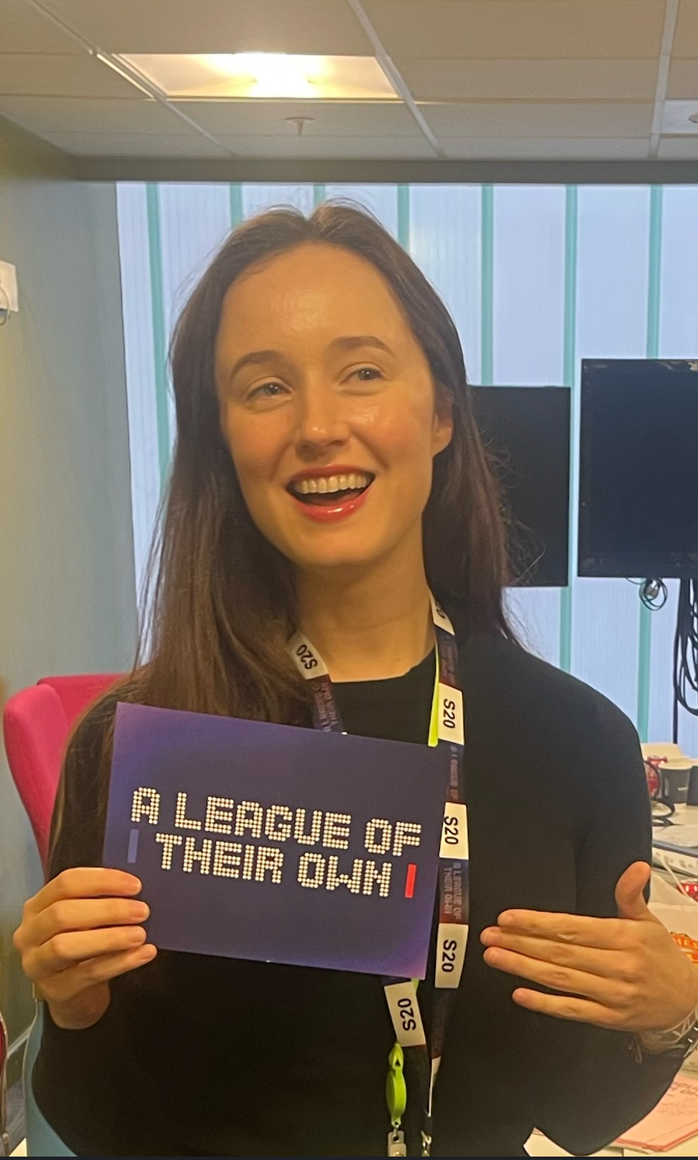 Katy Swainston, a woman with brown hair smiling, is holding a purple question card that reads 'A LEAGUE OF THEIR OWN' in a writers' room with large windows, monitors, and office supplies.