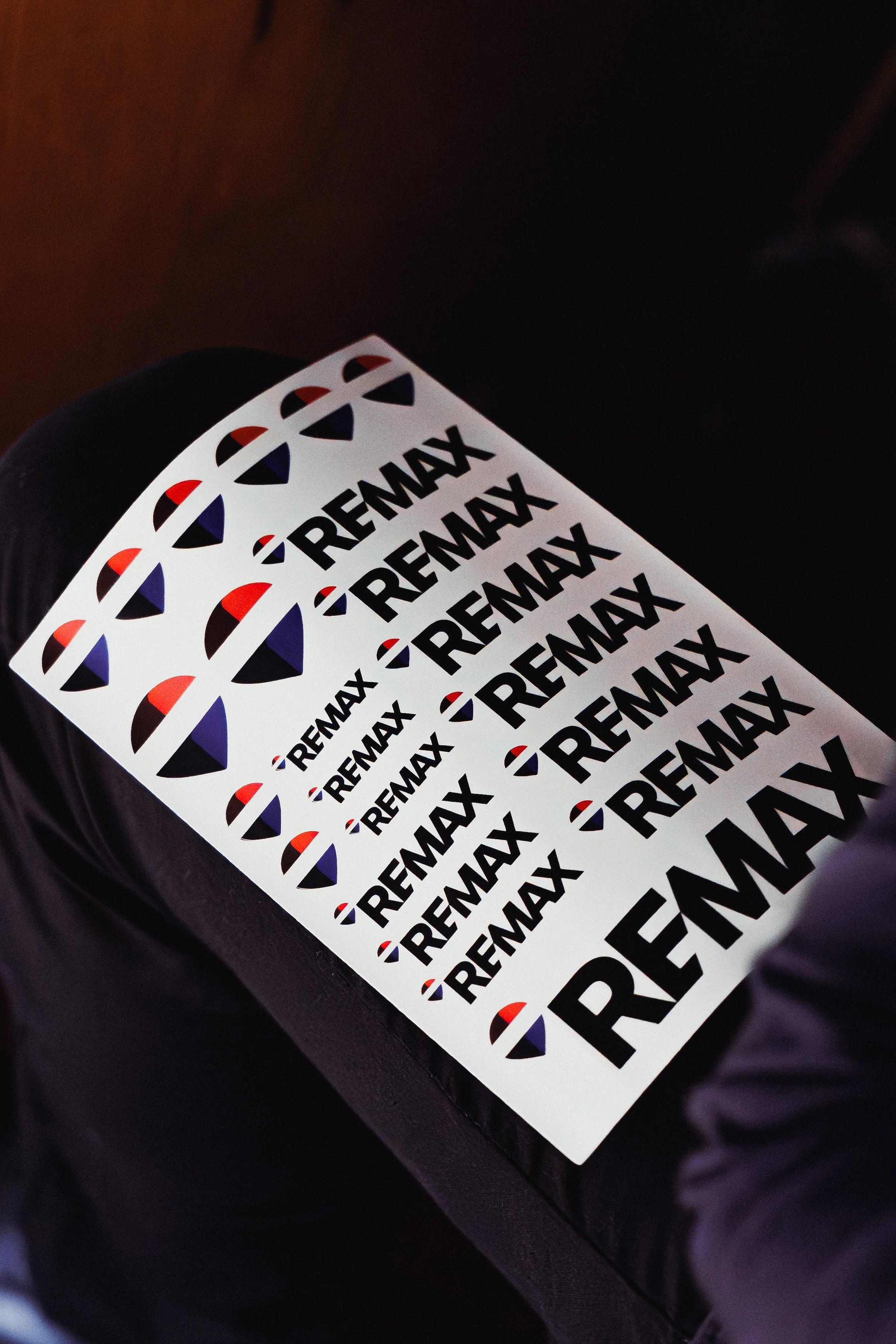 Multiple posters with the word 'REMAX' in black text on a white background, featuring the REMAX logo in red, white, and blue, placed on a black surface.