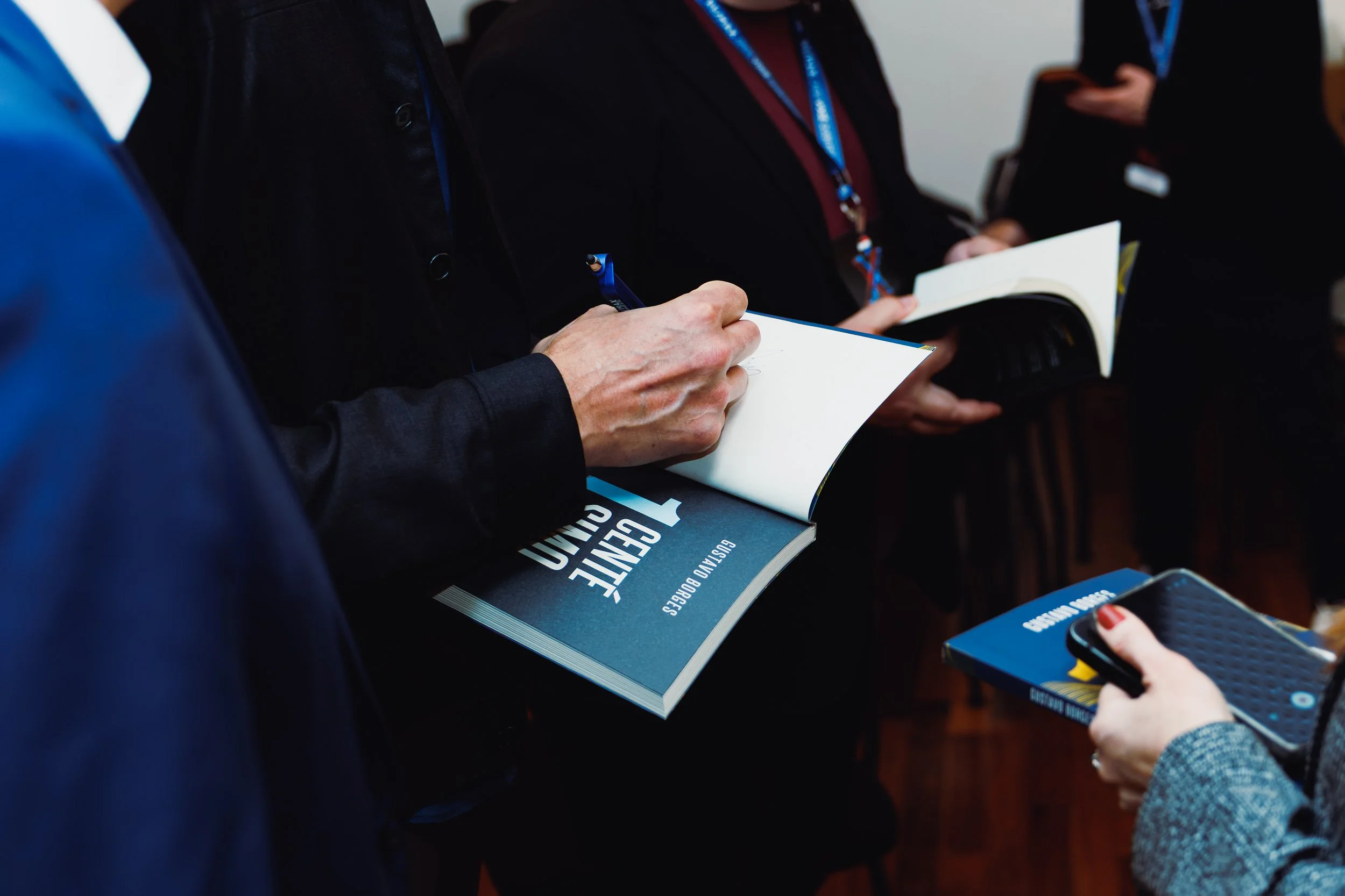 Close-up of people at a conference or seminar, some holding notebooks, pens, and pamphlets, with a focus on a person writing in a notebook.