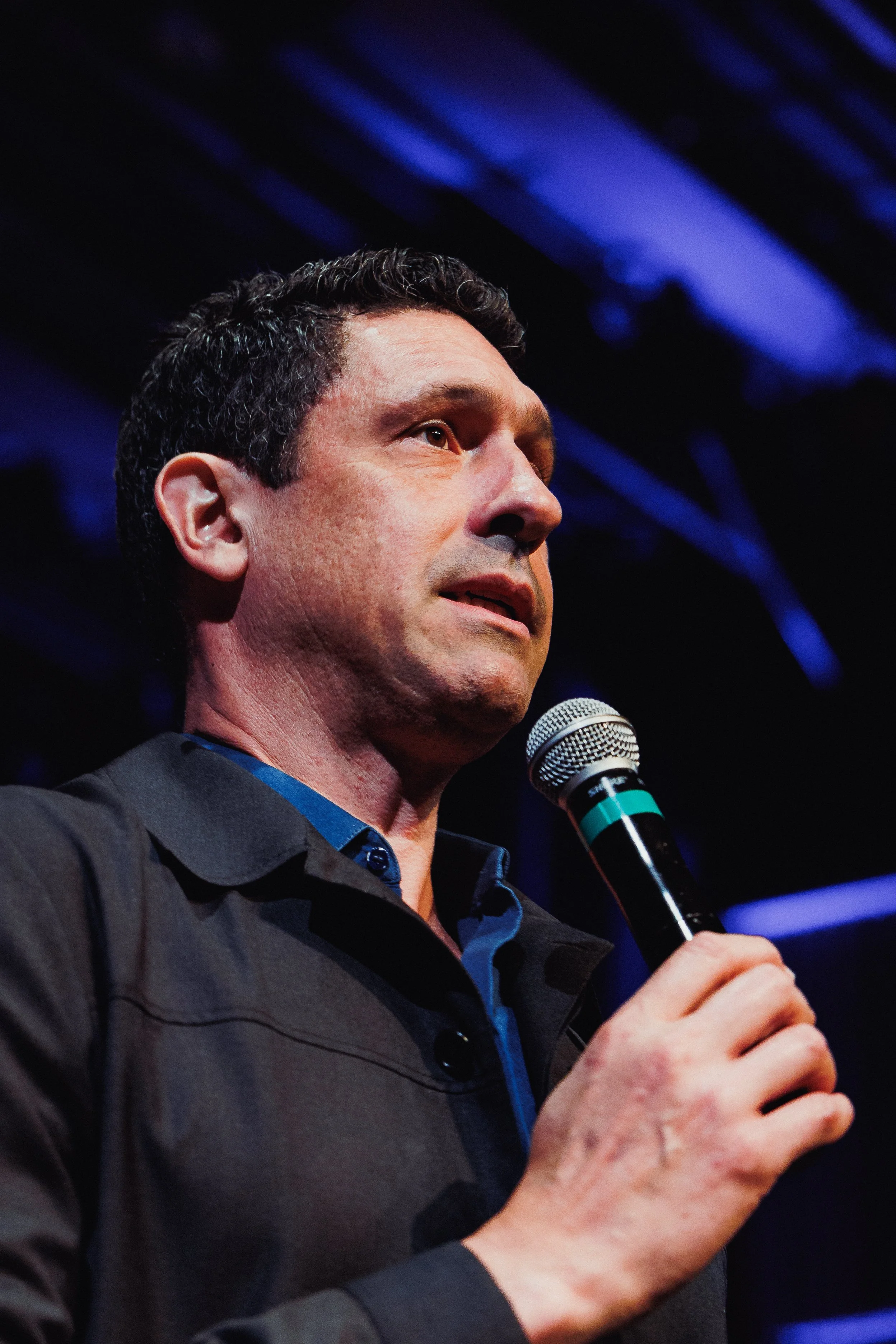 A man with dark, curly hair and light skin is speaking into a microphone. He is wearing a dark gray shirt with a blue collar, and the background features blue lighting.