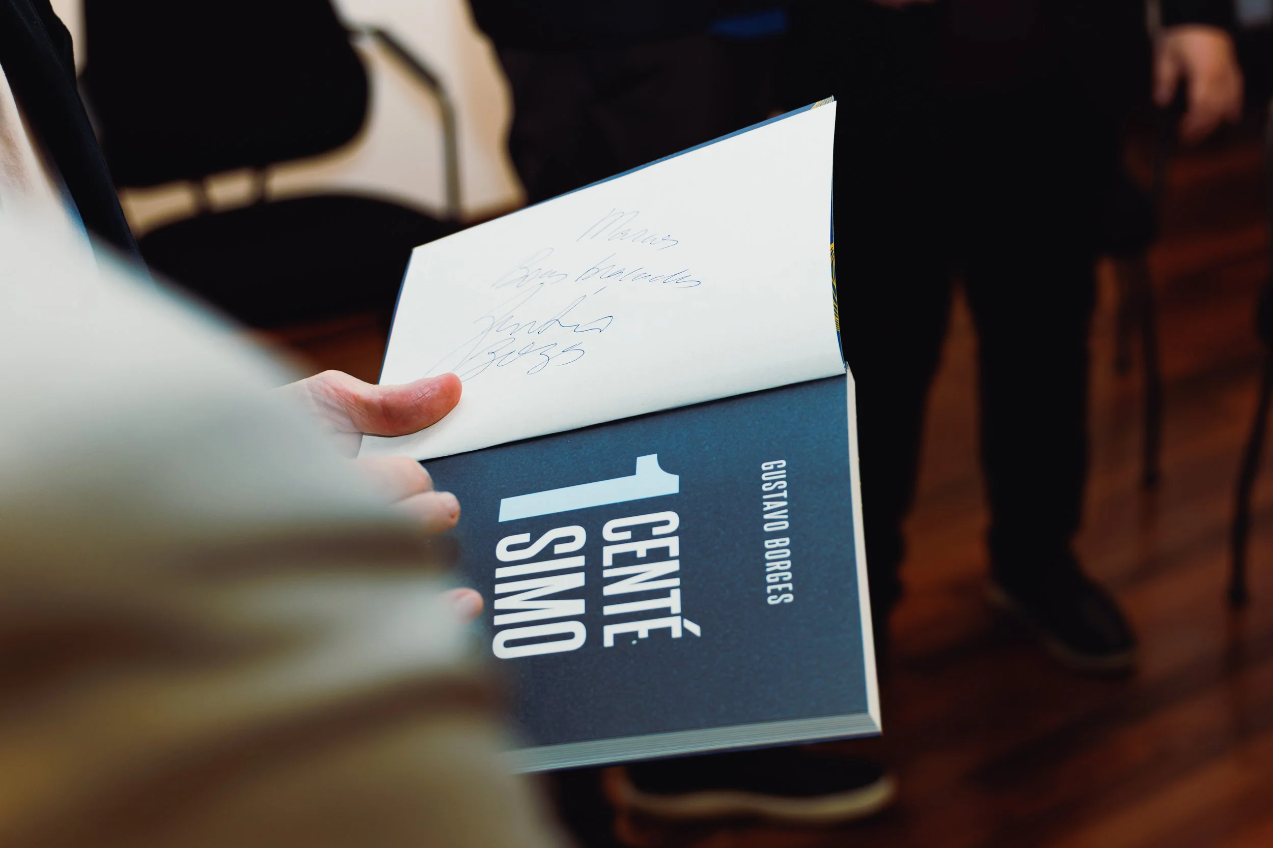 Person holding a brochure with handwritten notes on the front cover inside a room with chairs and people.