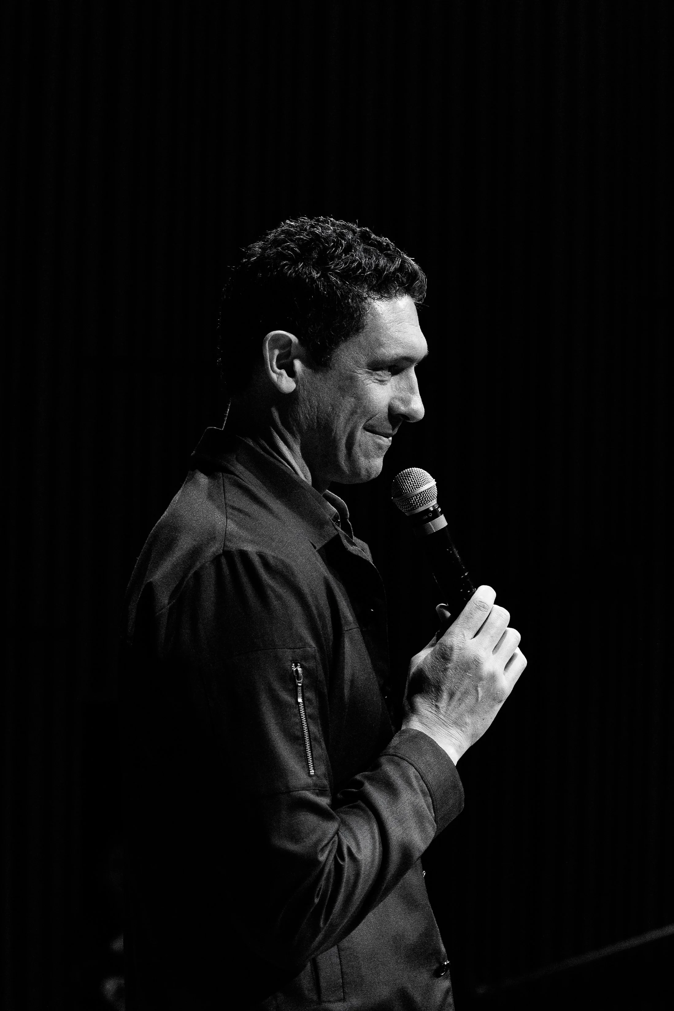 Black and white photo of a man holding a microphone and smiling, looking down, against a dark background.