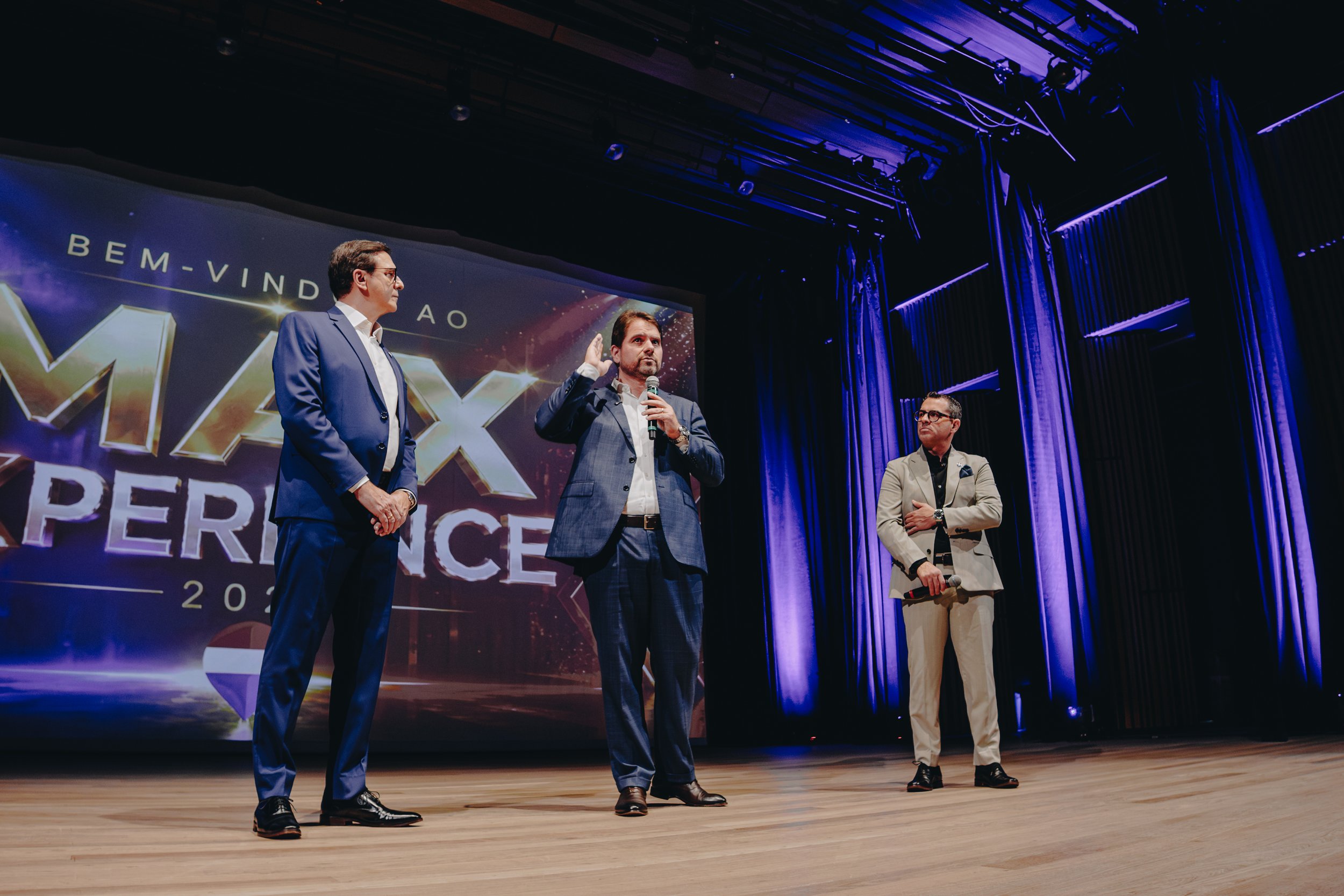 Three men on stage at a conference, dressed in suits, with one man speaking into a microphone. A large screen behind them displays the conference name, partially reading 'PERFORMANCE 2023'.