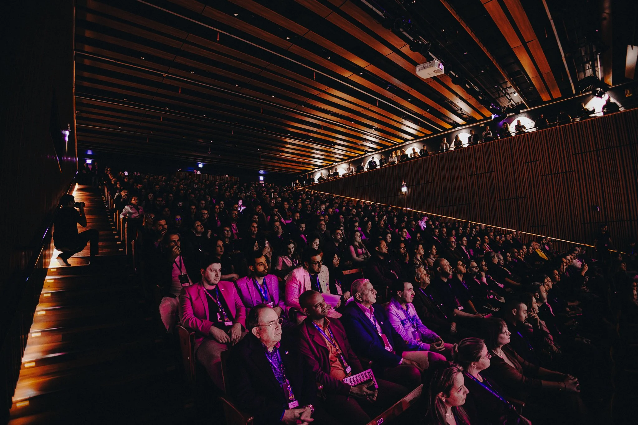 Audience seated in a dark auditorium, attending a conference or presentation, with some people on an upper balcony.