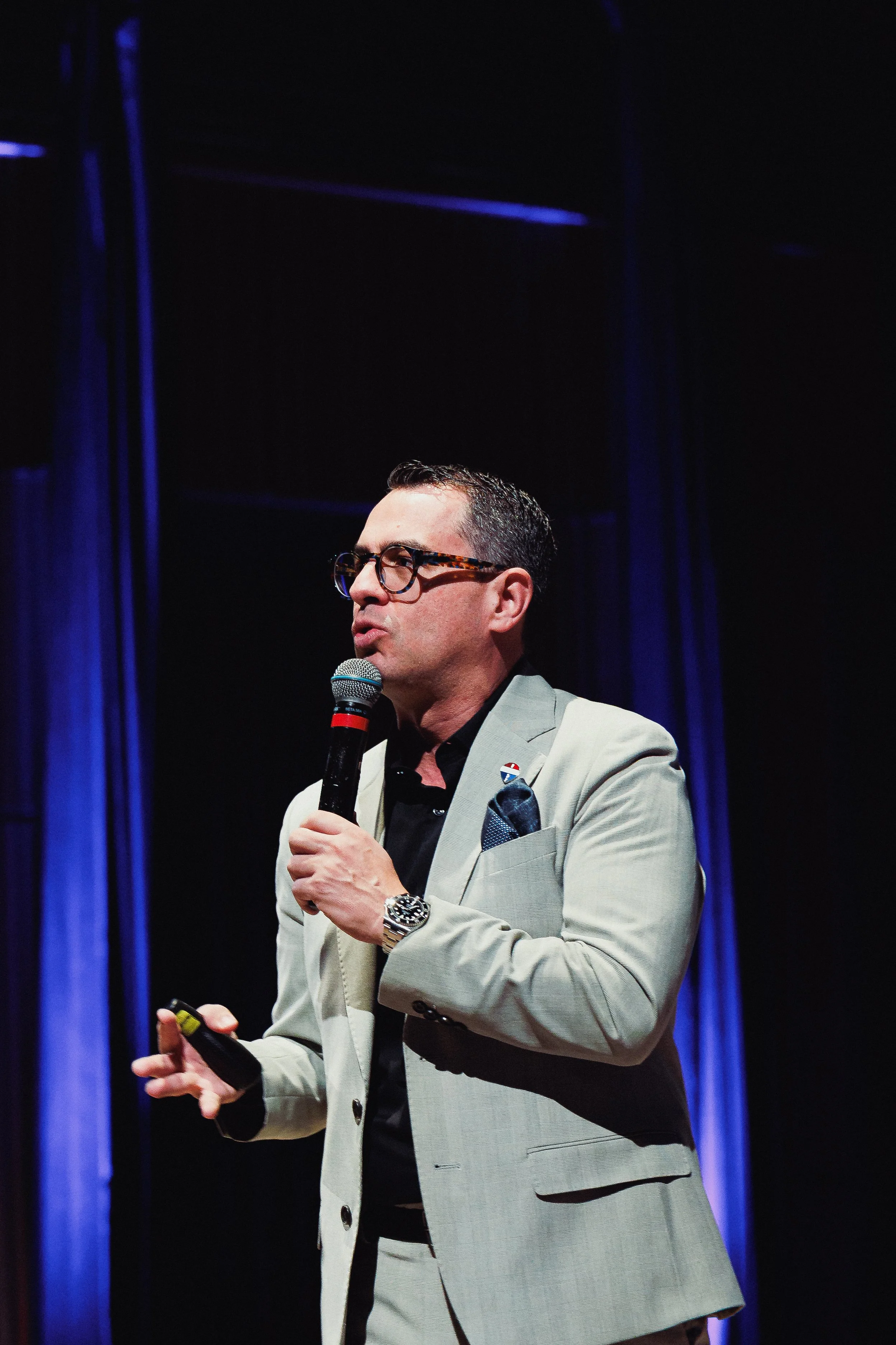 A man in a beige suit with glasses speaking into a microphone on stage, with dark background and blue lights.