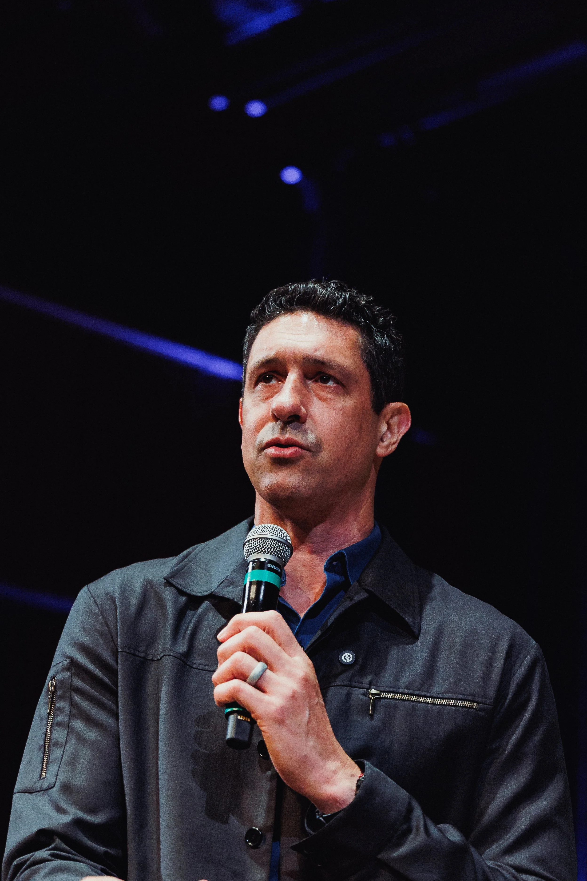 A man with short dark hair holding a microphone, dressed in a dark jacket and a blue shirt, speaking or presenting on stage in a dark setting with purple lights above.