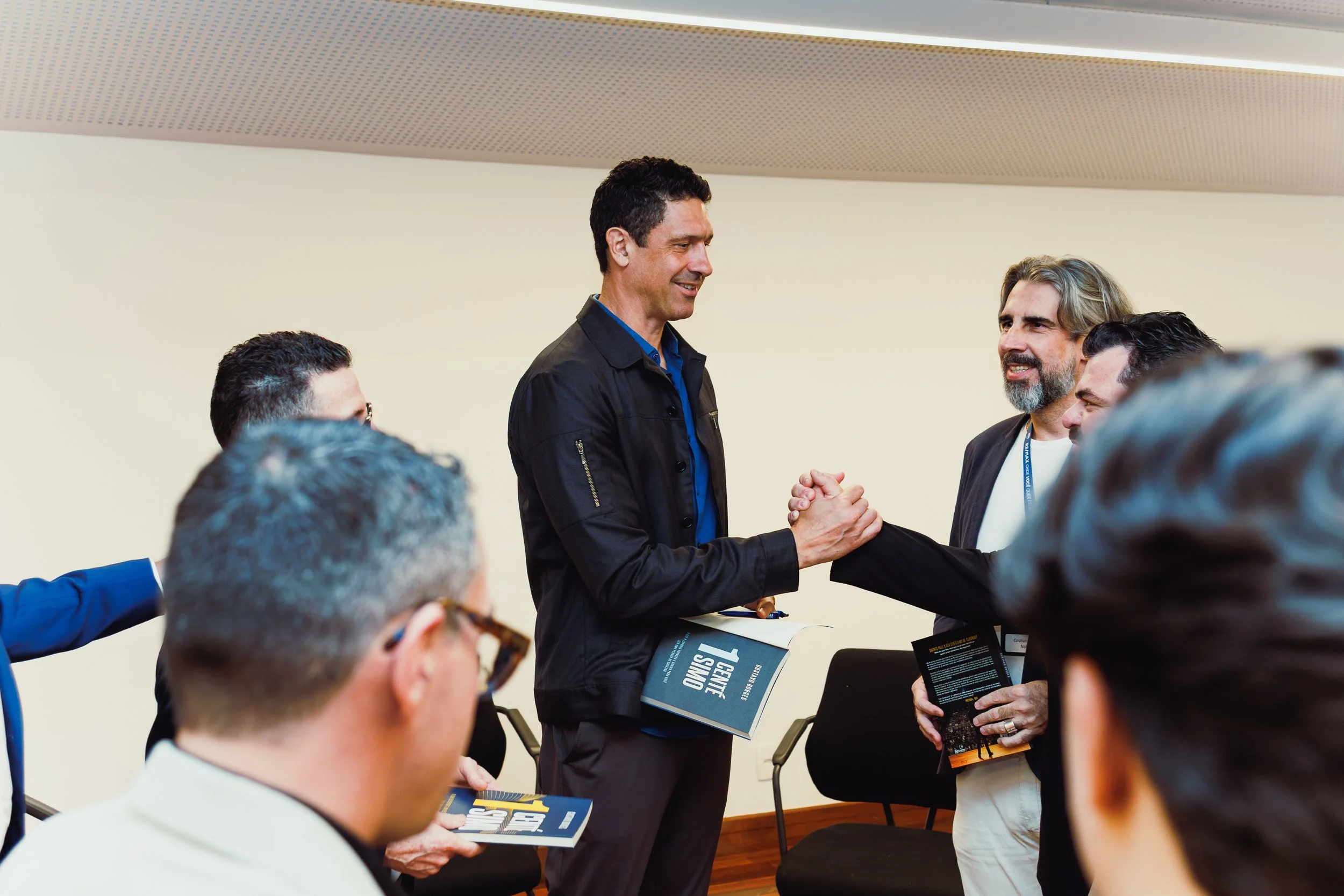 Two men shaking hands in a professional setting, with multiple people observing. One man has dark hair and is wearing a black jacket, the other has gray hair and a beard, both dressed in business casual attire. They appear to be engaging in a friendl