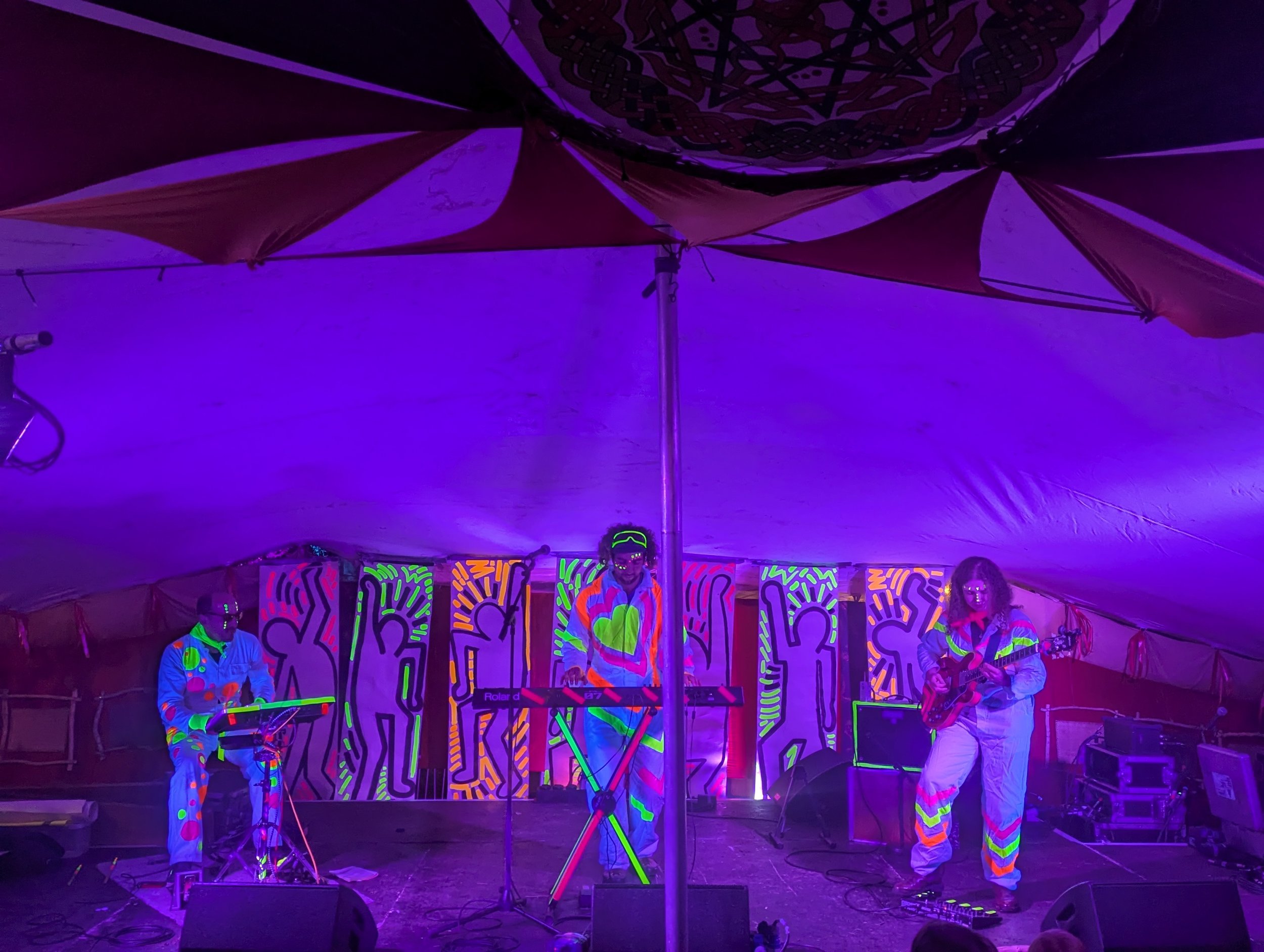ChokKa performs at a festival with neon-colored clothing and body paint, on a stage decorated with vibrant, psychedelic patterns.