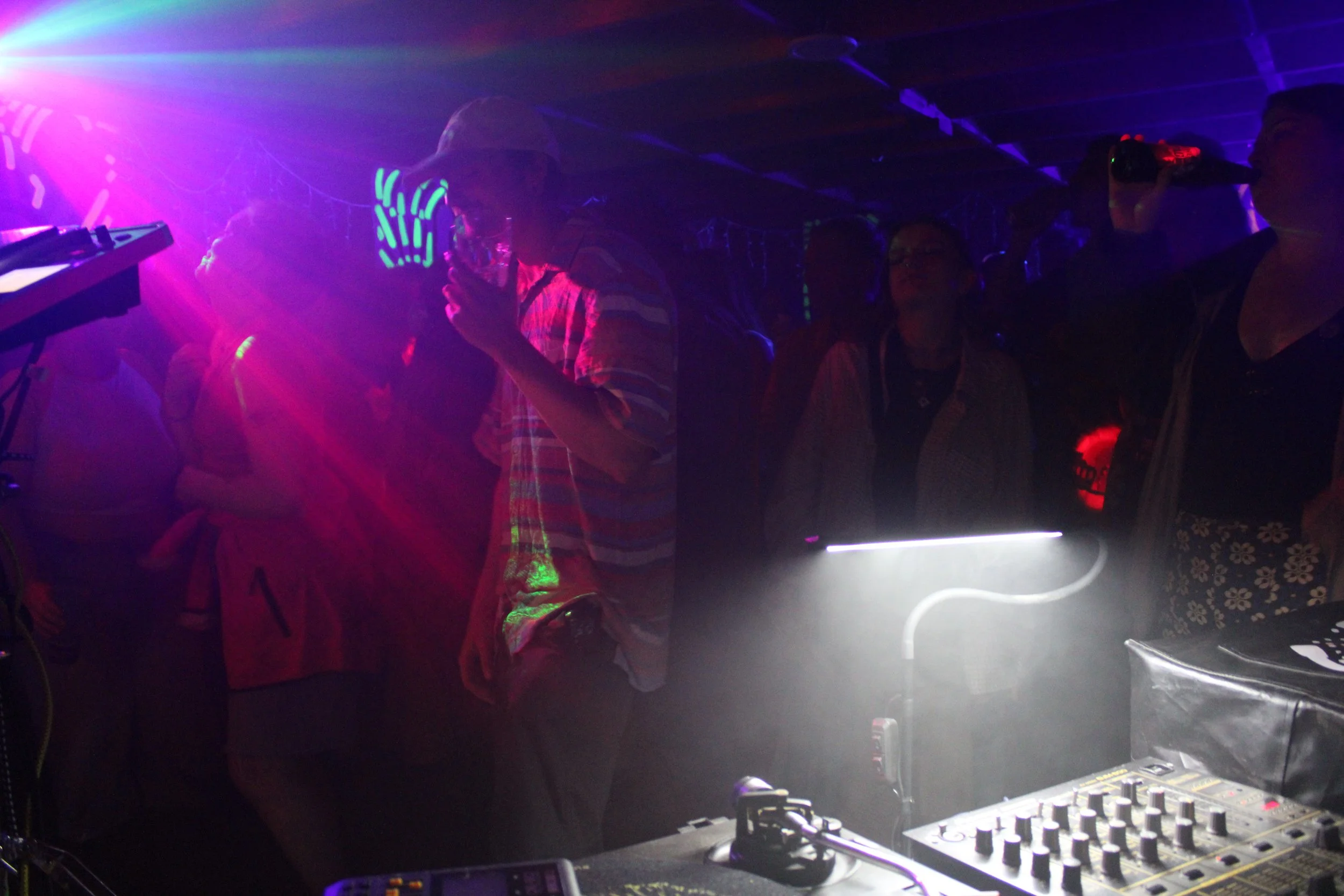 People dancing at a ChokKa party with colorful lights, a DJ booth, and a person taking a picture or video with a phone.
