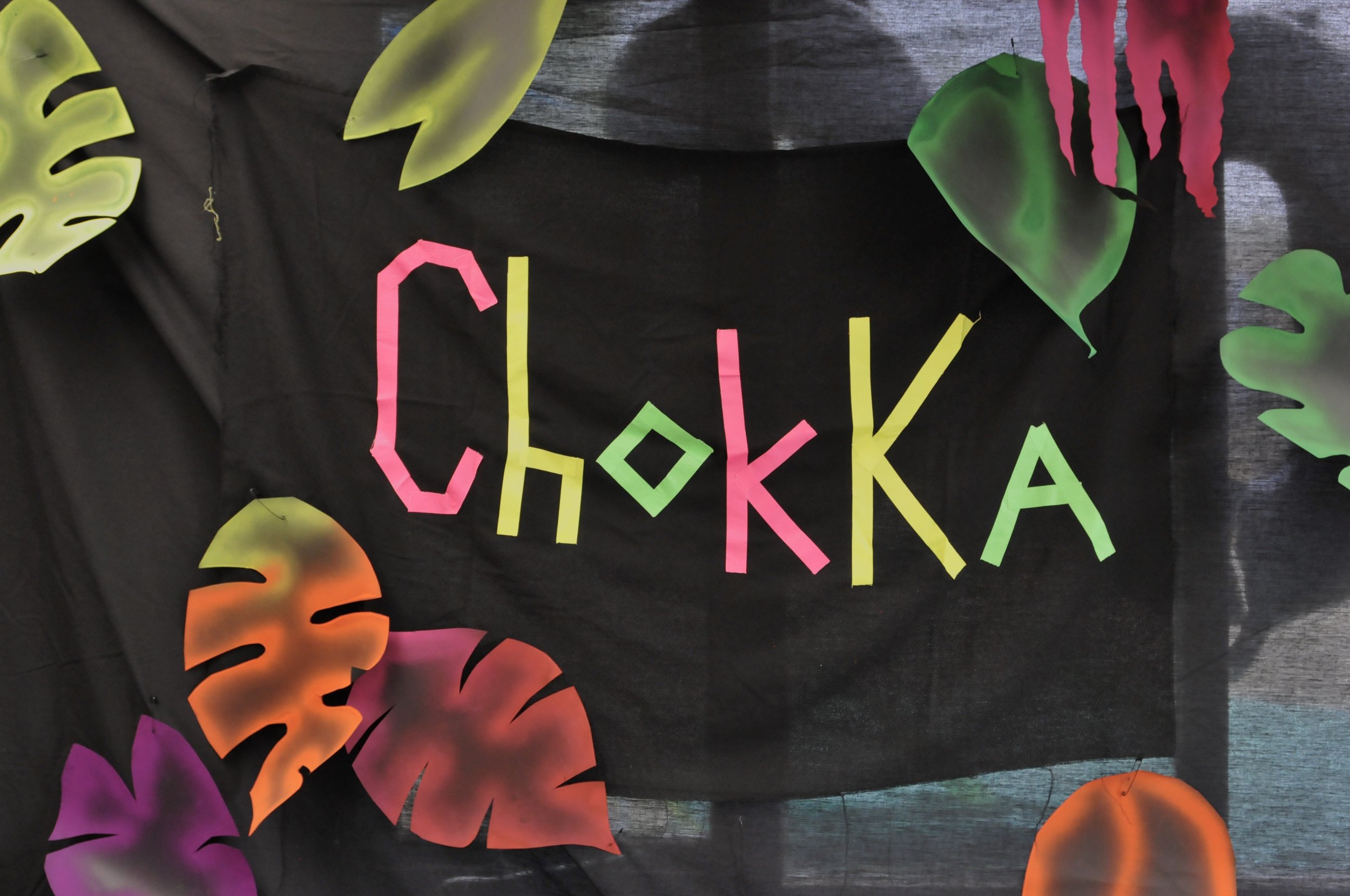 Colorful paper cutouts of tropical leaves and the word 'Checka' written in colorful paper letters on a black background.