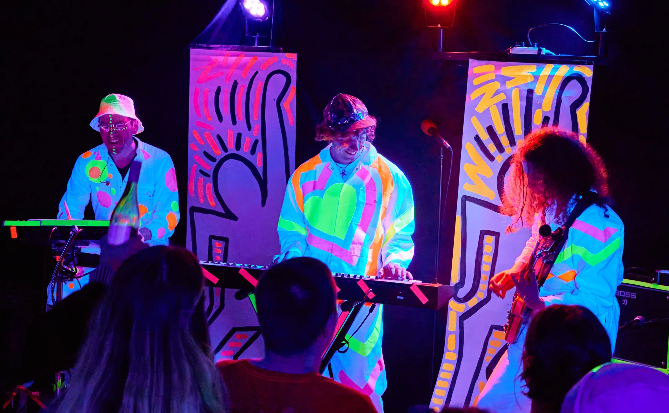 A colorful neon-lit music performance with three musicians in vibrant, glow-in-the-dark outfits on stage, surrounded by bright, abstract banners.