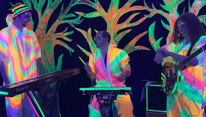 Three musicians wearing colorful, glowing outfits perform on stage with a backdrop of neon painted trees, playing electronic keyboard, electronic drums, and guitar.