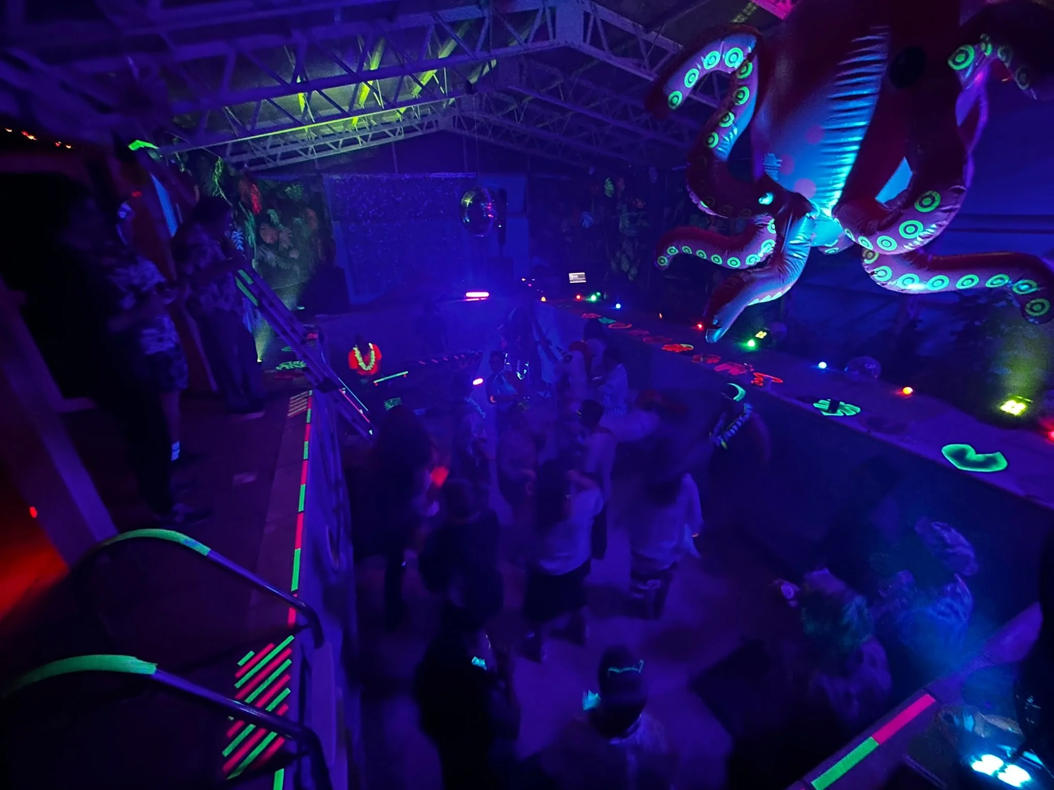 People at the ChokKa pool party with large inflatable octopus and balloon-like decorations, illuminated by blacklight with neon colors.