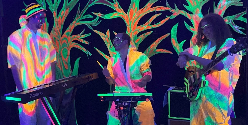 Three band members performing music with colorful glow-in-the-dark body paint and neon clothing, in front of a vibrant, painted backdrop of trees, with one playing a keyboard, one playing drums, and one playing an electric guitar.