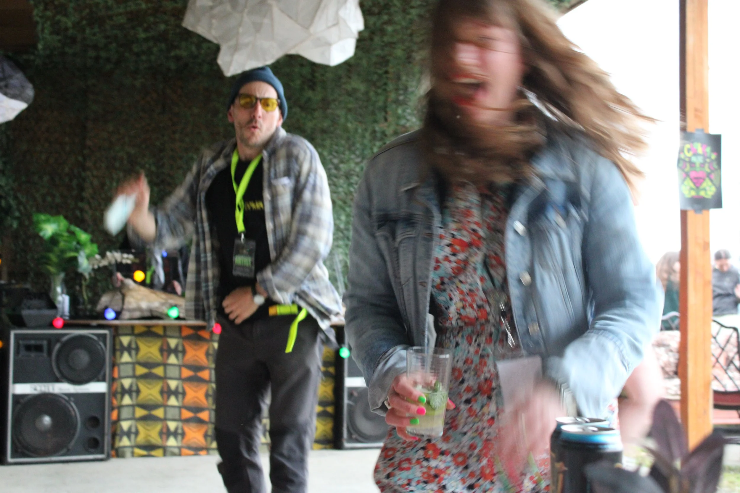 Two women and a man at a social gathering. One man is focused, wearing sunglasses and a beanie, standing near a DJ setup with speakers. One woman in the foreground has curly hair and is holding a drink, smiling with her mouth open. Another woman in t