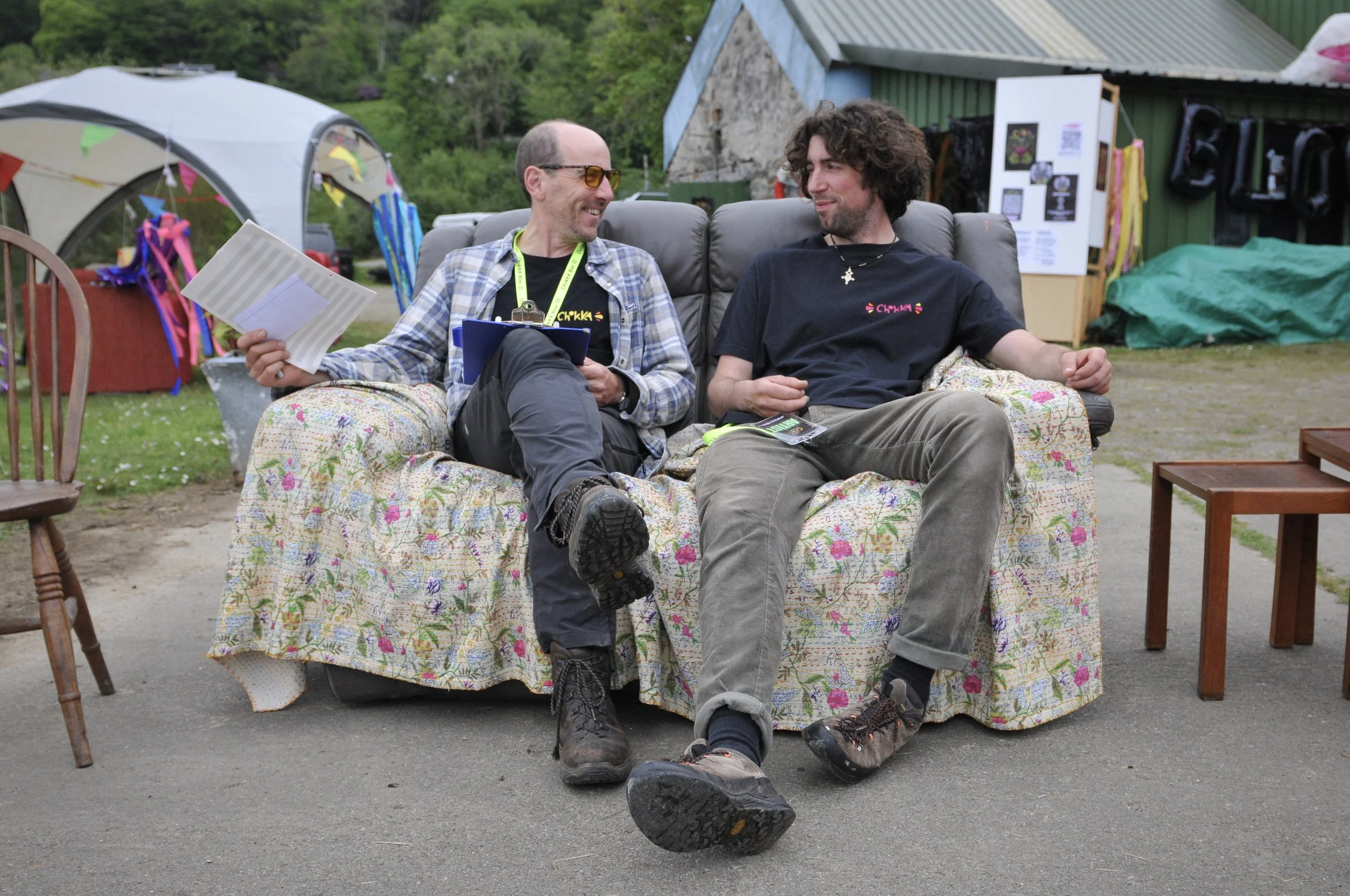 Two men sitting on a floral patterned sofa outdoors, smiling at each other. One man is wearing glasses, a plaid shirt, and holding papers, while the other wears a black t-shirt with a logo, has curly hair, and a necklace. There are colorful decoratio