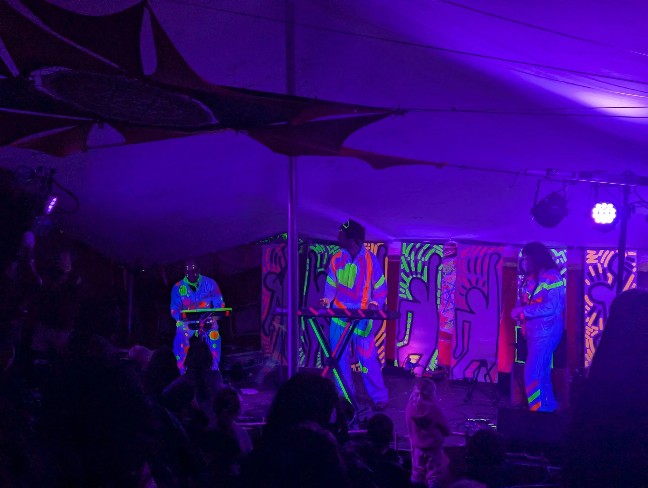 A group of musicians performing at a night event with black lights, wearing outfits with neon patterns, and a colorful graffiti backdrop.