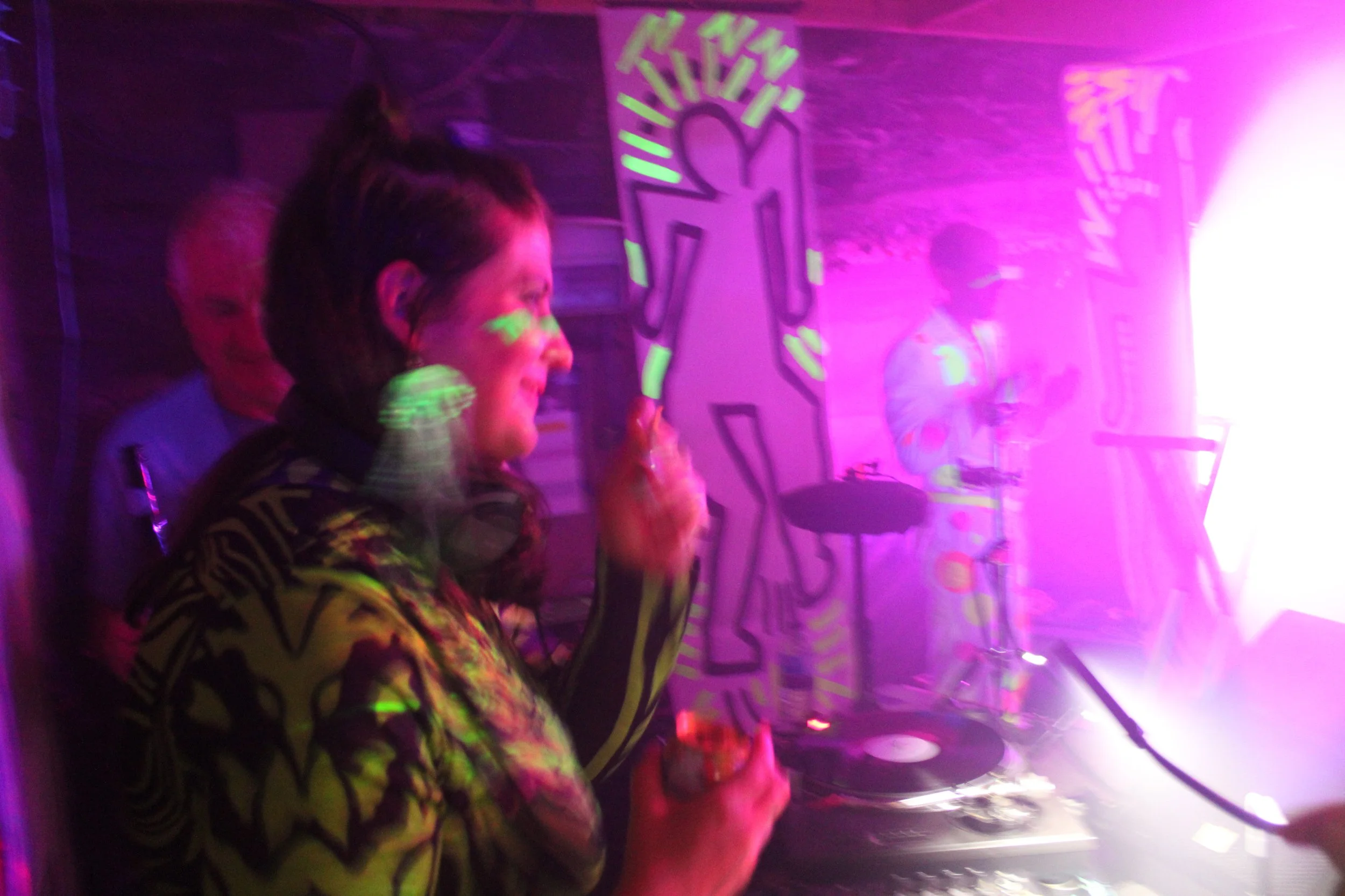 A woman with pink and purple lighting in a room with colorful graffiti art, holding what appears to be a orange drink, possibly at a party or club.