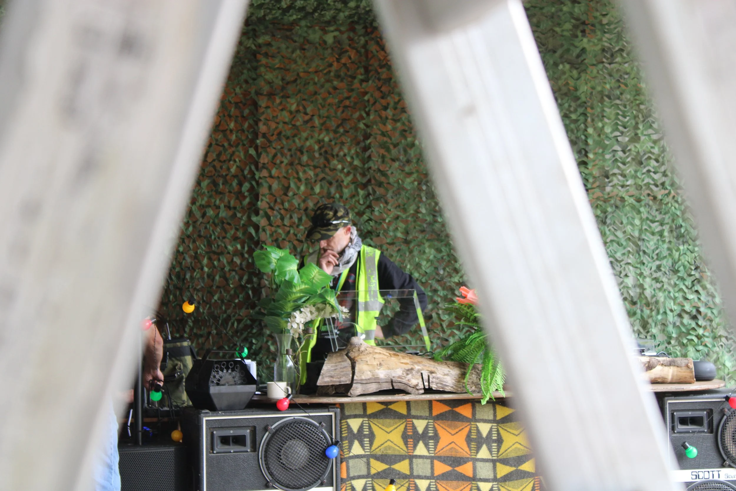 A man wearing glasses, a yellow reflective vest, and a black hat is standing behind a DJ station with electronic equipment and a log, in a room with a camo-themed wall and plants. The photo is taken through a frame made of metallic bars with colorful