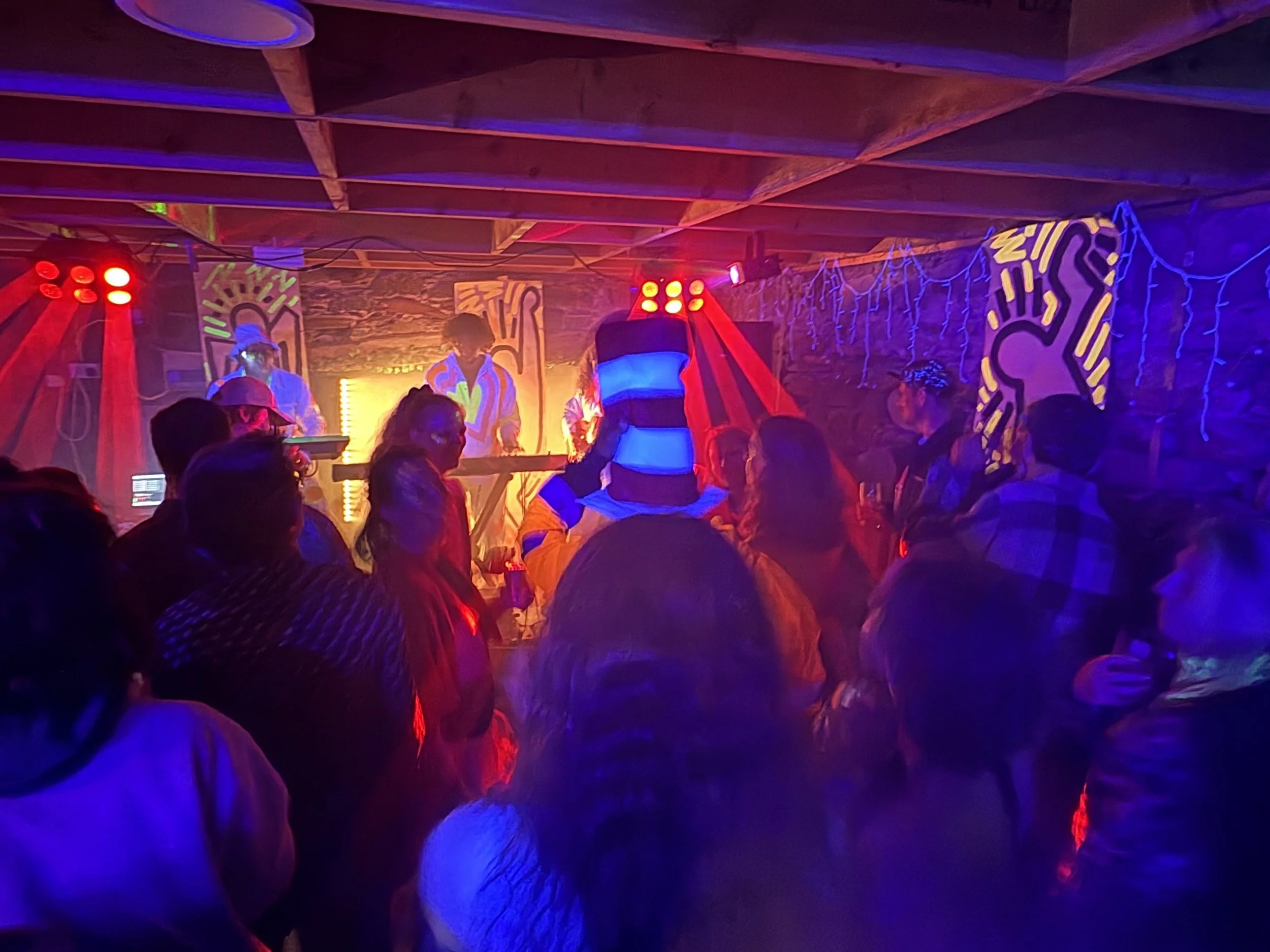 A dimly lit nightclub or party scene with colorful neon and LED lighting, people dancing, and abstract wall art with vibrant colors and shapes.