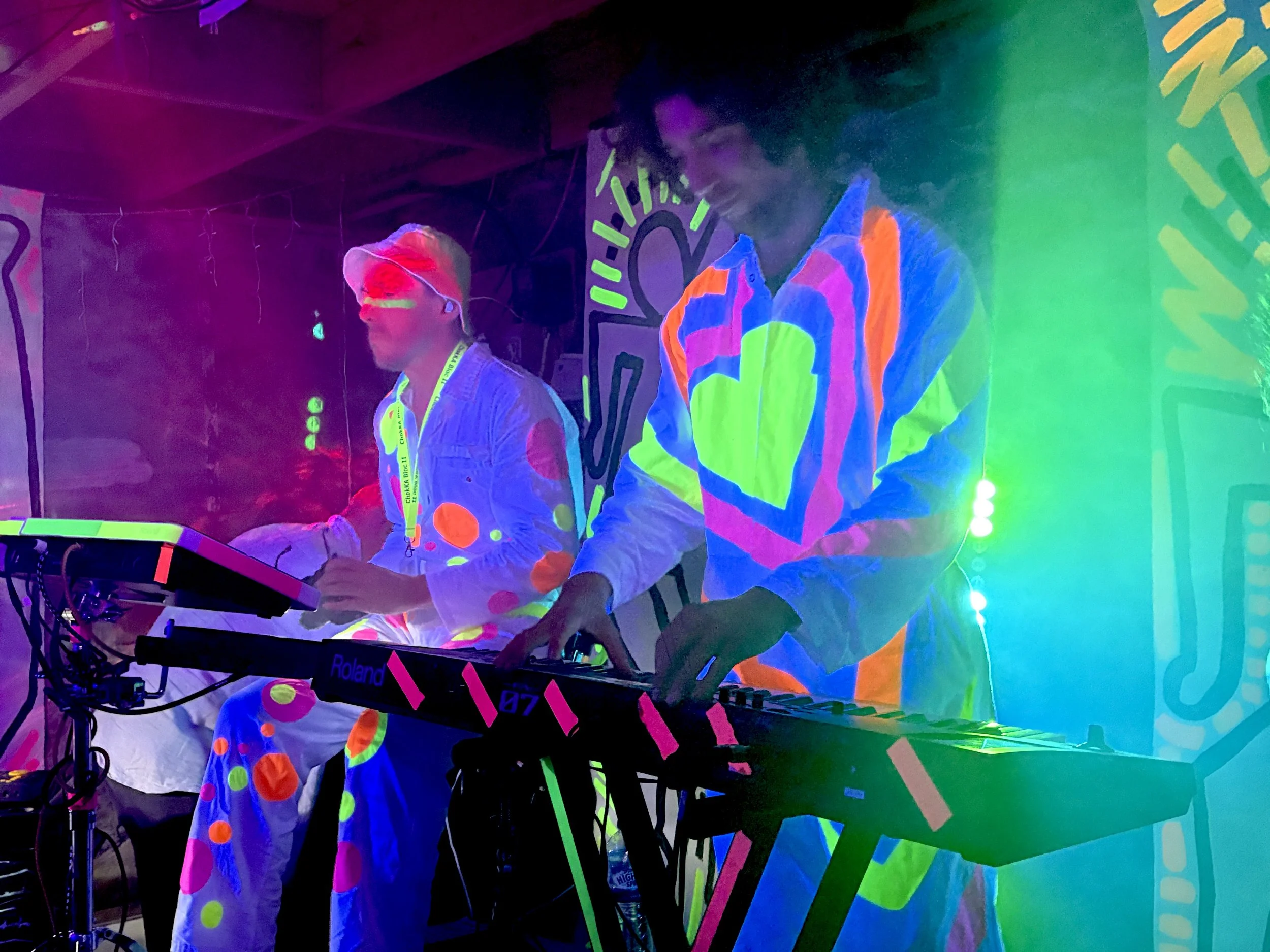 Two individuals playing electronic music on a synthesizer in a dark room illuminated by blacklight and colorful, neon decorations.