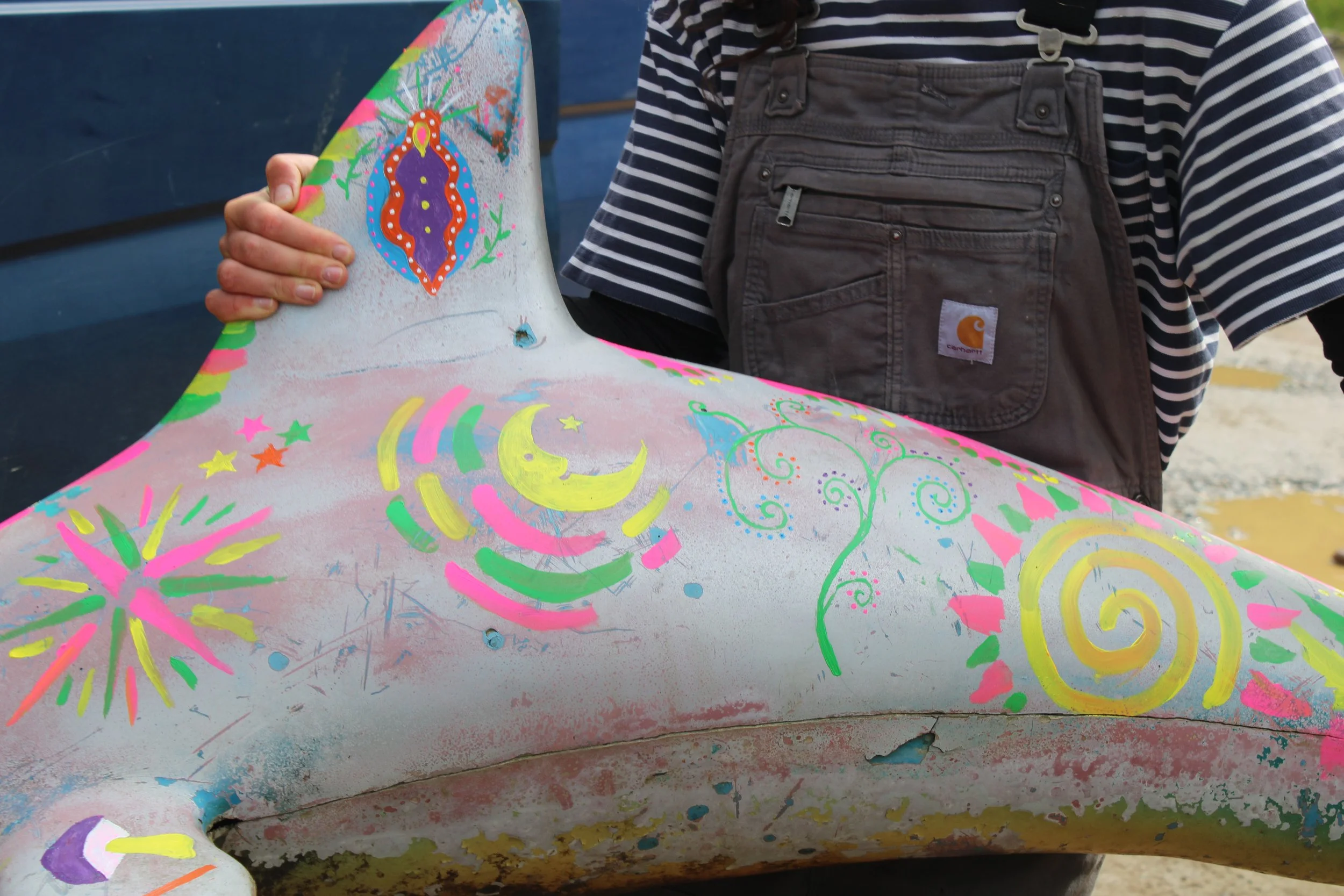 Close-up of a colorful painted surface with fireworks, stars, moons, and swirling patterns. Part of a person's striped shirt and brown overalls are visible, holding the painted surface.