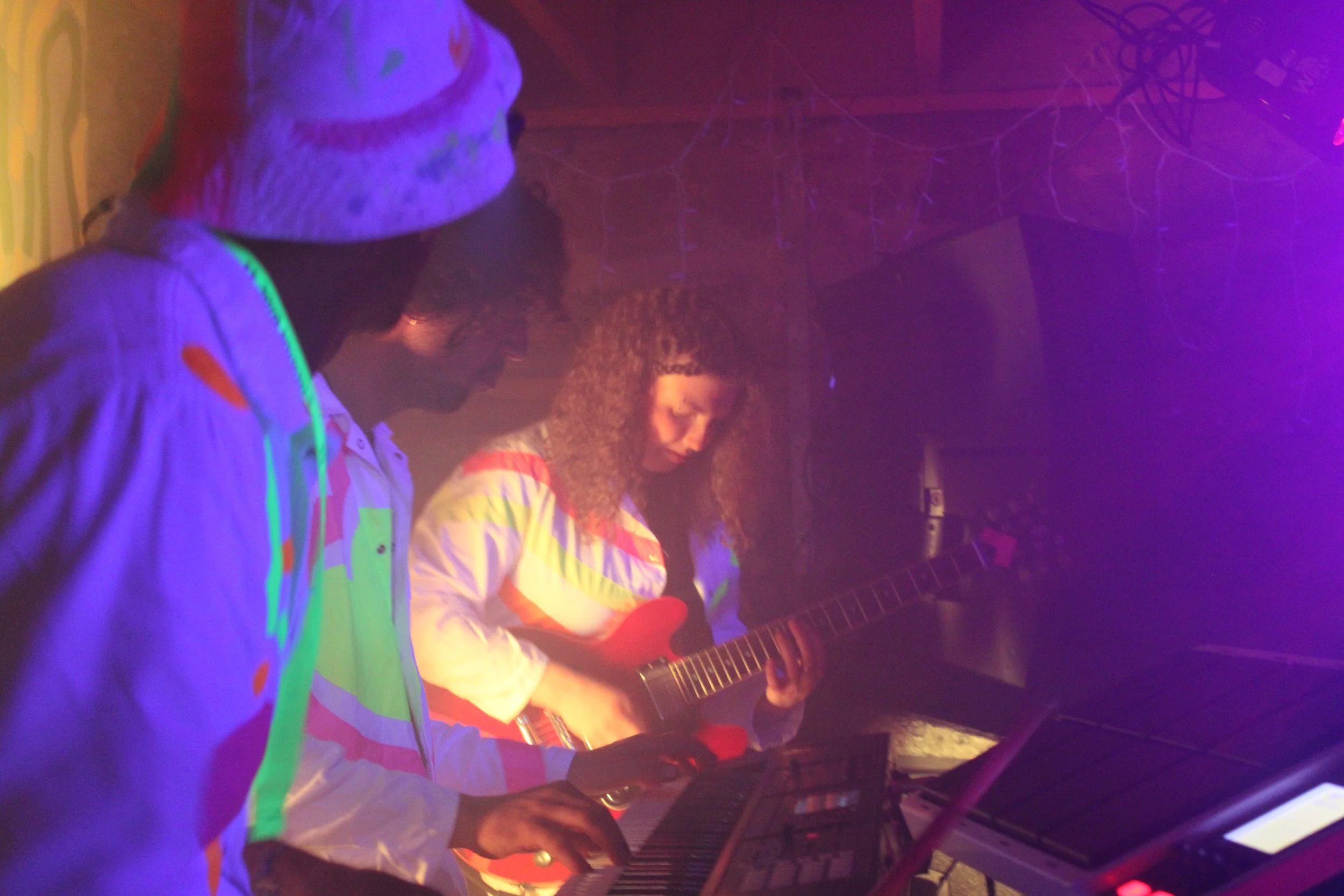 Two people play electric guitar in a dimly lit room with colorful lighting.