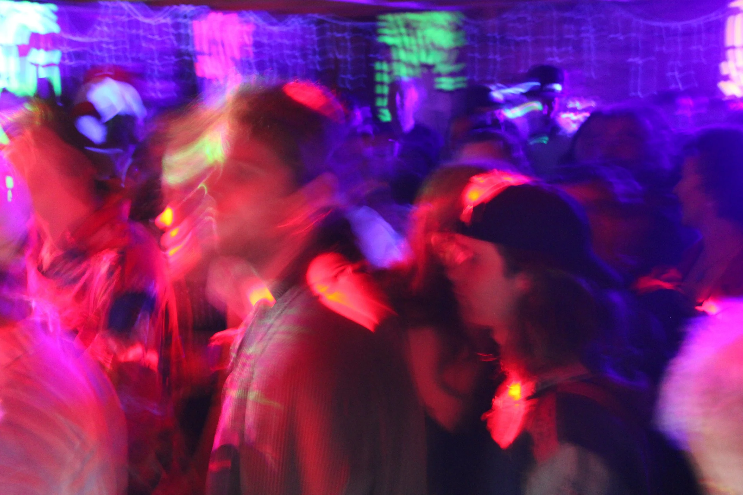 Blurred image of a crowd dancing in a dimly lit space with colorful lighting, mainly purple, red, and green.