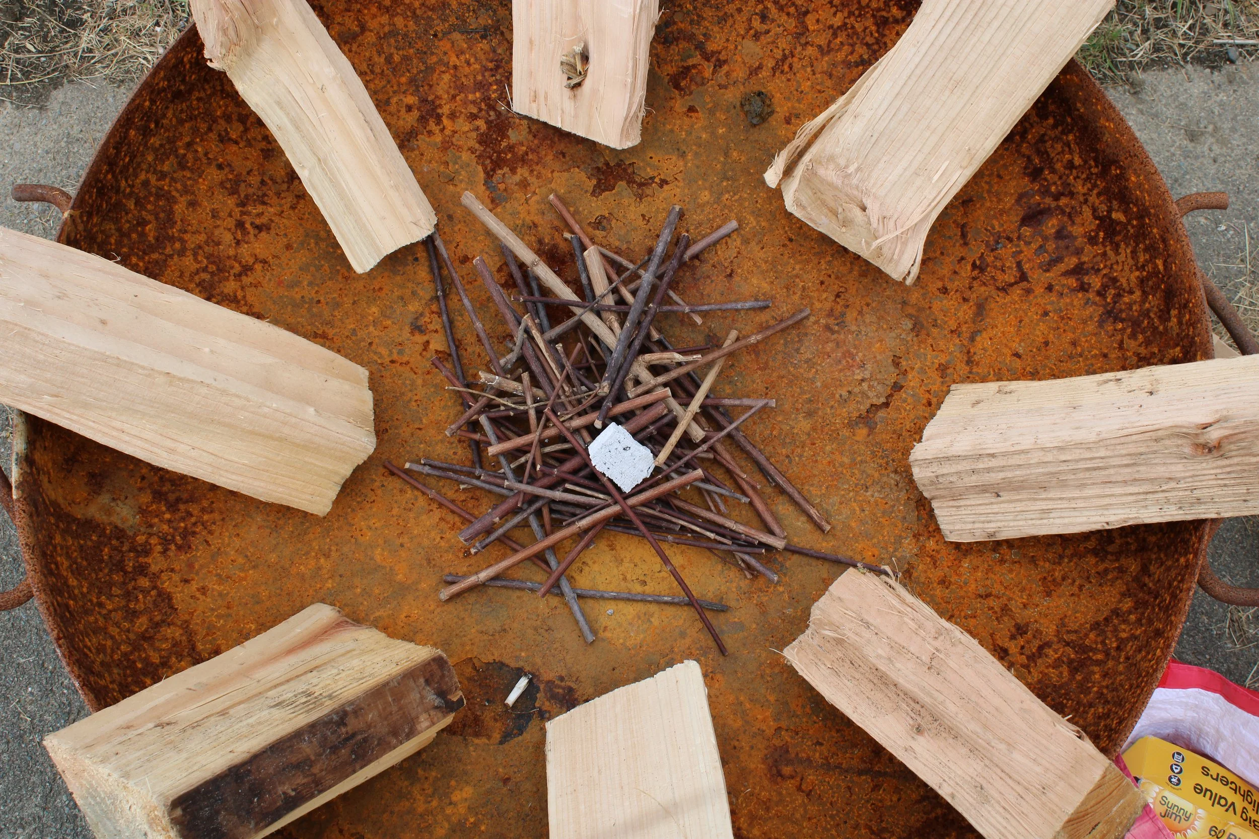A rusted fire pit with large pieces of split wood arranged around the edge and a pile of small sticks and nails in the center.