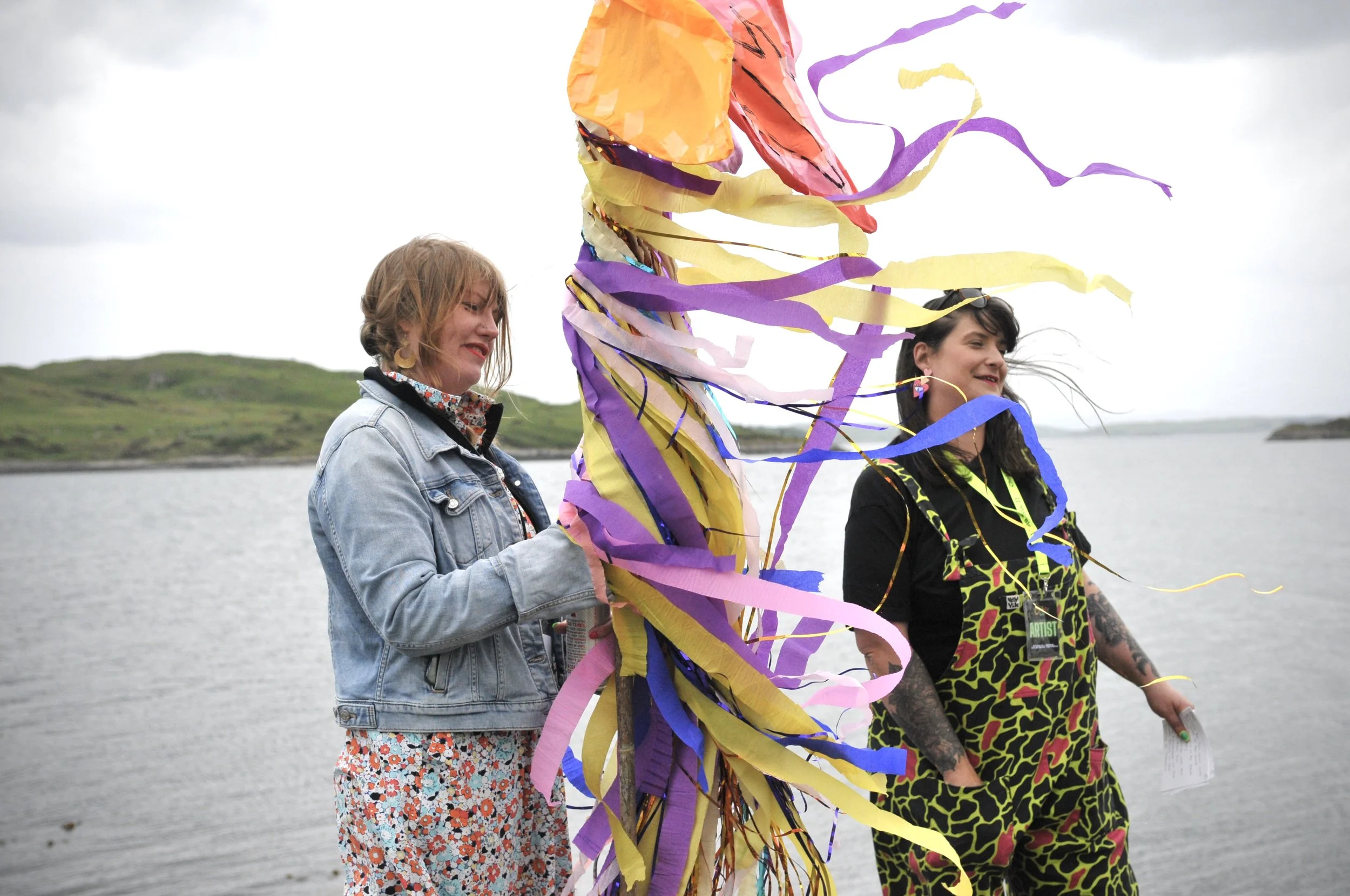 Two women on a beach with a colorful kite. One woman with blonde hair in a denim jacket and floral dress holds a soda can, while the other with dark hair and tattoos wears a neon camouflage outfit. The sky is cloudy, and grassy hills are visible in the background.