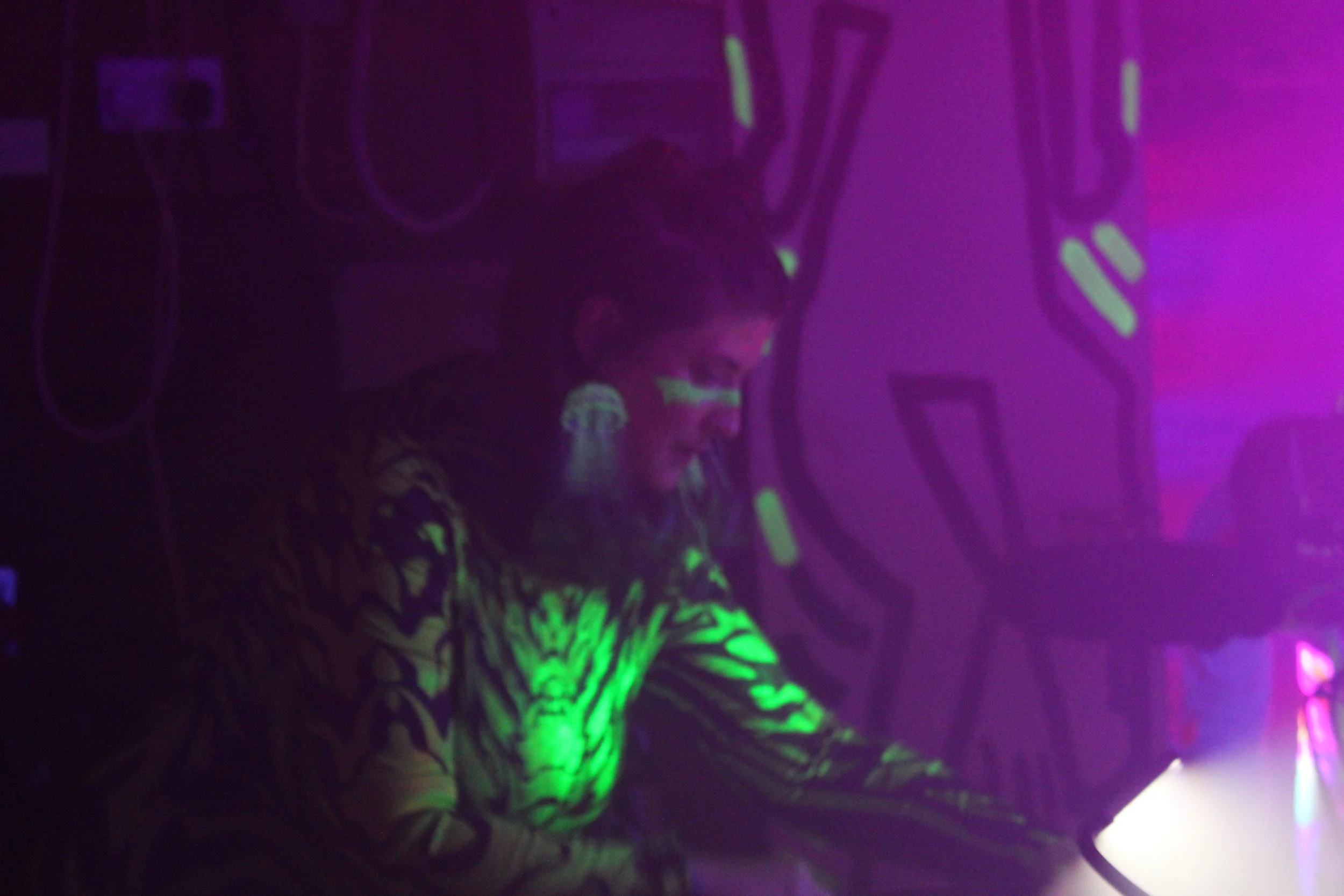 A person wearing neon green and black clothing with neon green patterns, sitting in a dark room illuminated by blacklight, making parts of their outfit glow in the dark. The background is dark with some light-colored objects and wires visible.