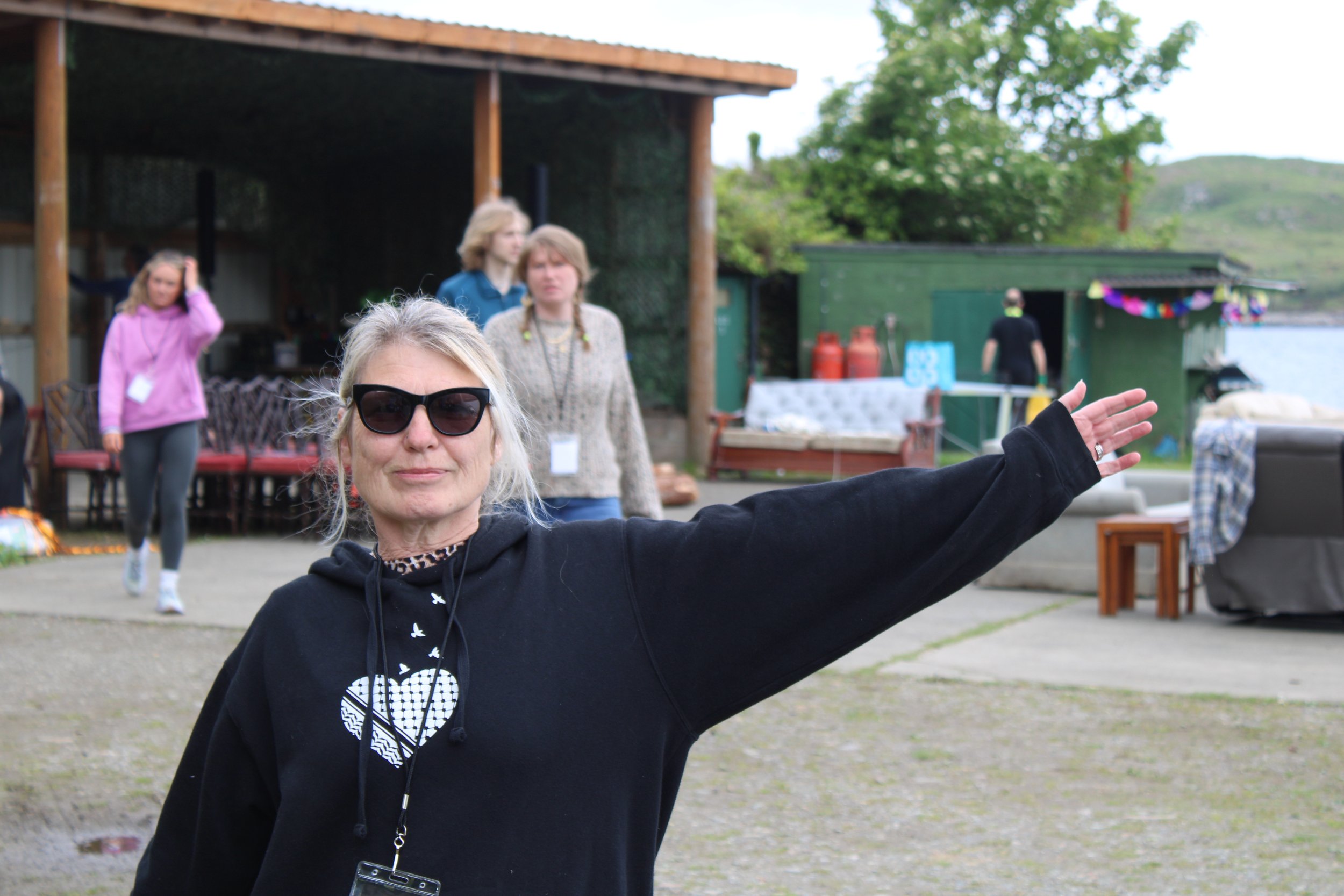 A woman wearing sunglasses and a black hoodie extending her arm, standing outdoors with a group of people and a wooden structure in the background.