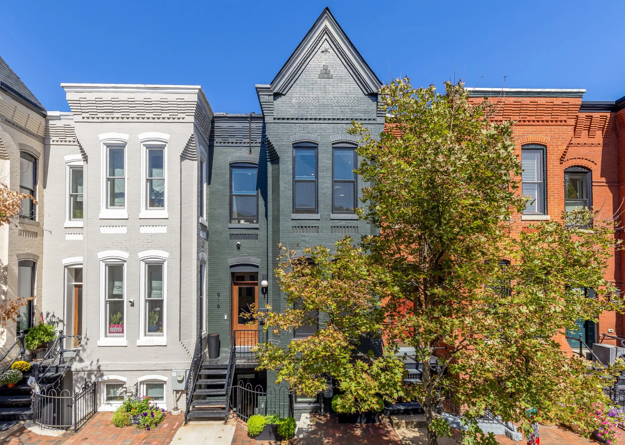 A row of colorful Victorian-style houses, each painted in white, dark gray, and red brick, with a tree in front partially obscuring the buildings and a clear blue sky above.