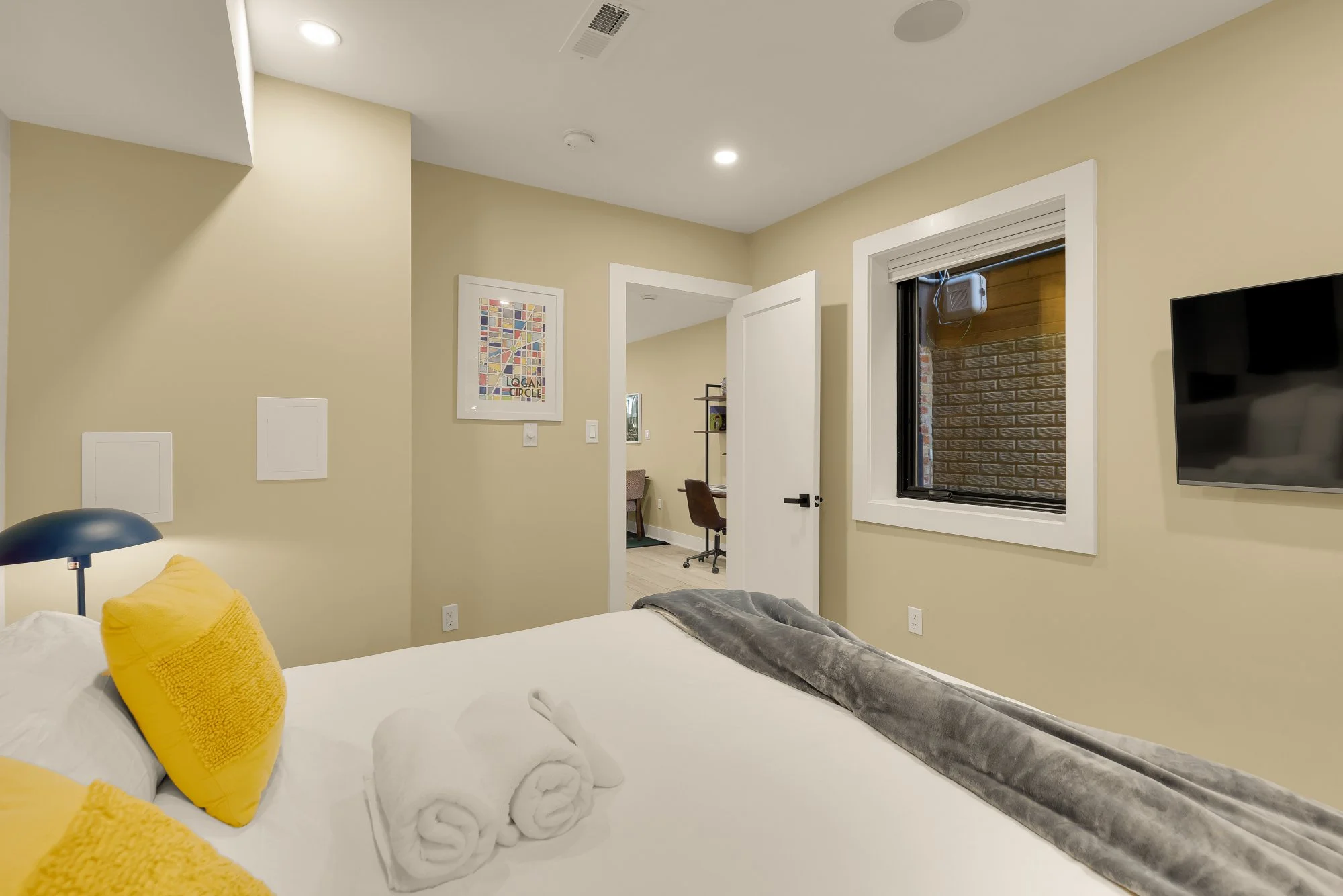 Inside a bedroom, a bed with yellow pillows and white towels folded at the foot. The wall has a colorful, geometric artwork and a window with an exterior brick wall visible. Part of a desk and office chair can be seen through a doorway.