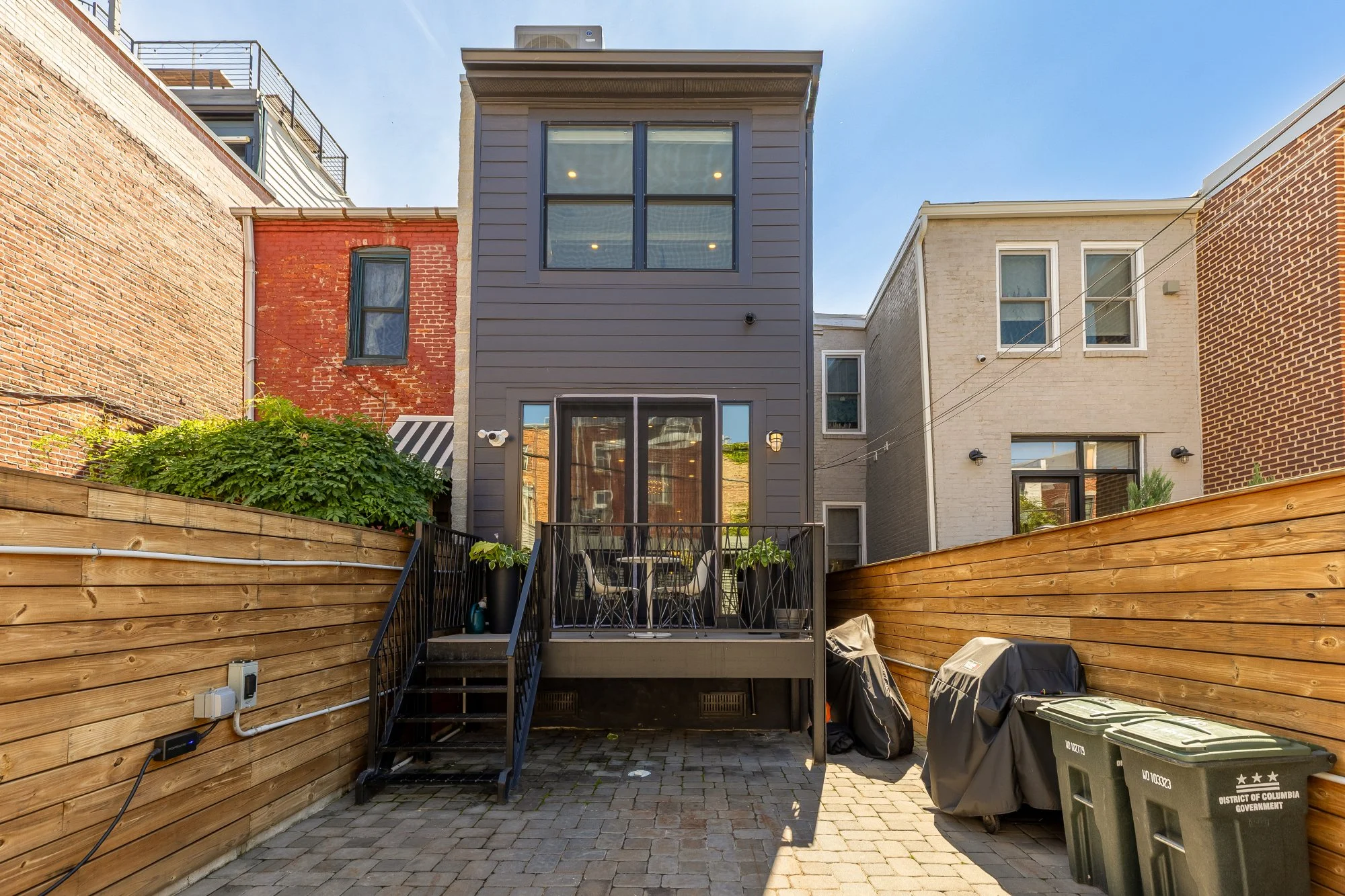 Backyard of a modern house with a raised deck and stairs, surrounded by a wooden privacy fence, with neighboring brick buildings on either side, and covered with cobblestone pavement.