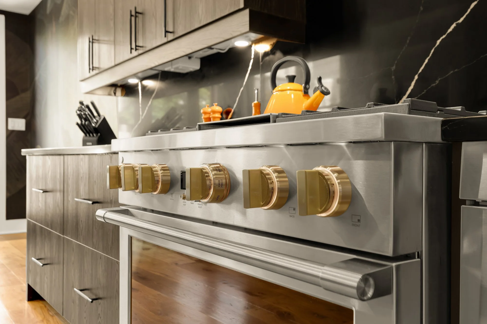 Modern kitchen with stainless steel stove with yellow control knobs, a yellow teapot on the burner, and black knife set on the counter. Wooden cabinets are above, and black marble backsplash. Wooden floor and ambient lighting.