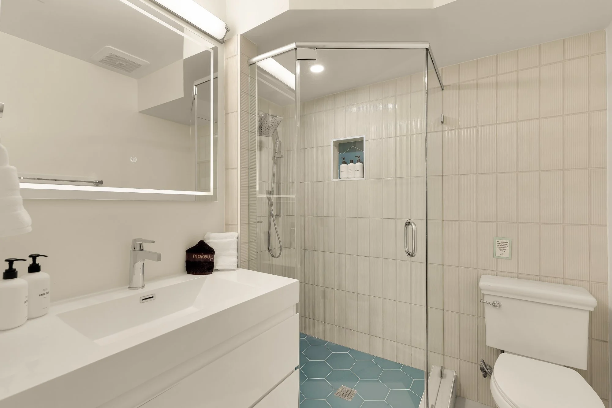 Modern bathroom featuring a white sink with a faucet, bottles of hand soap, a mounted mirror, a cubicle with a glass door and showerhead, beige tiled walls, a toilet, and blue hexagonal floor tiles.