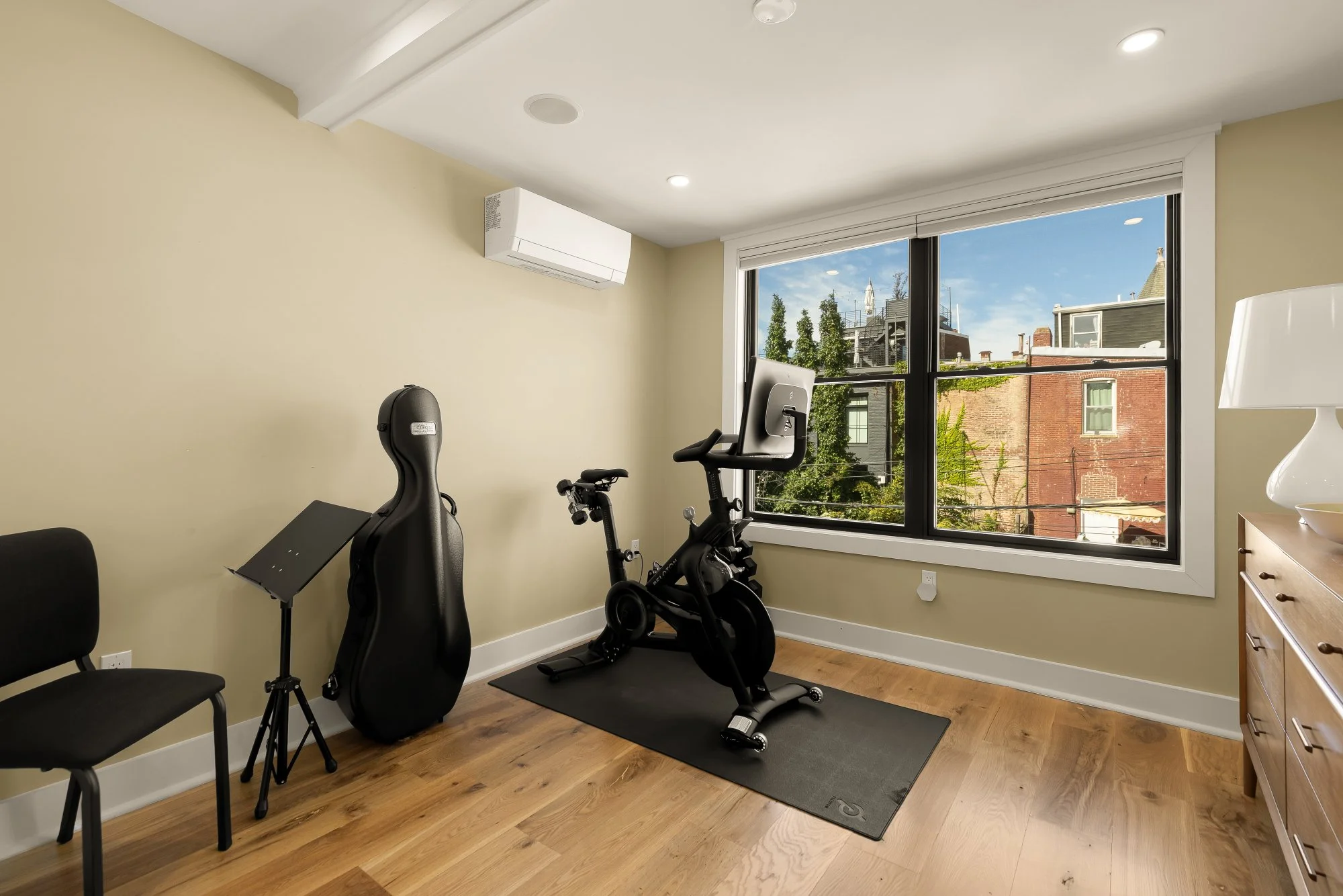 A bright home gym with a large window showing neighboring buildings and trees outside. The room has light-colored walls, wooden flooring, and ceiling lights. Fitness equipment includes an exercise bike, elliptical trainer, and a black guitar case. A 