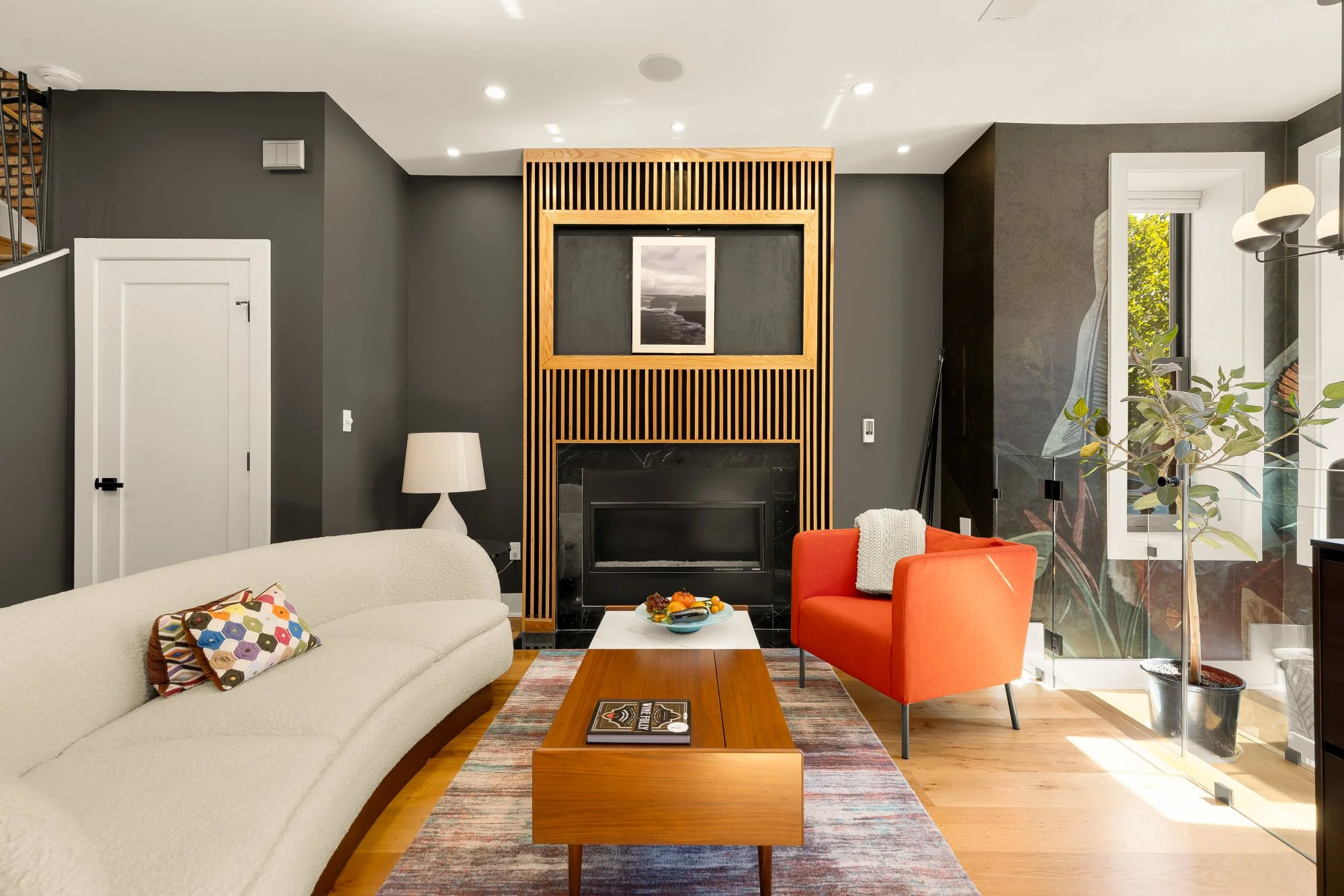 Modern living room with a beige sofa, a wooden coffee table, two orange armchairs, a white table lamp, a fireplace with a black marble surround, and a large window with greenery outside. The walls are painted dark gray.