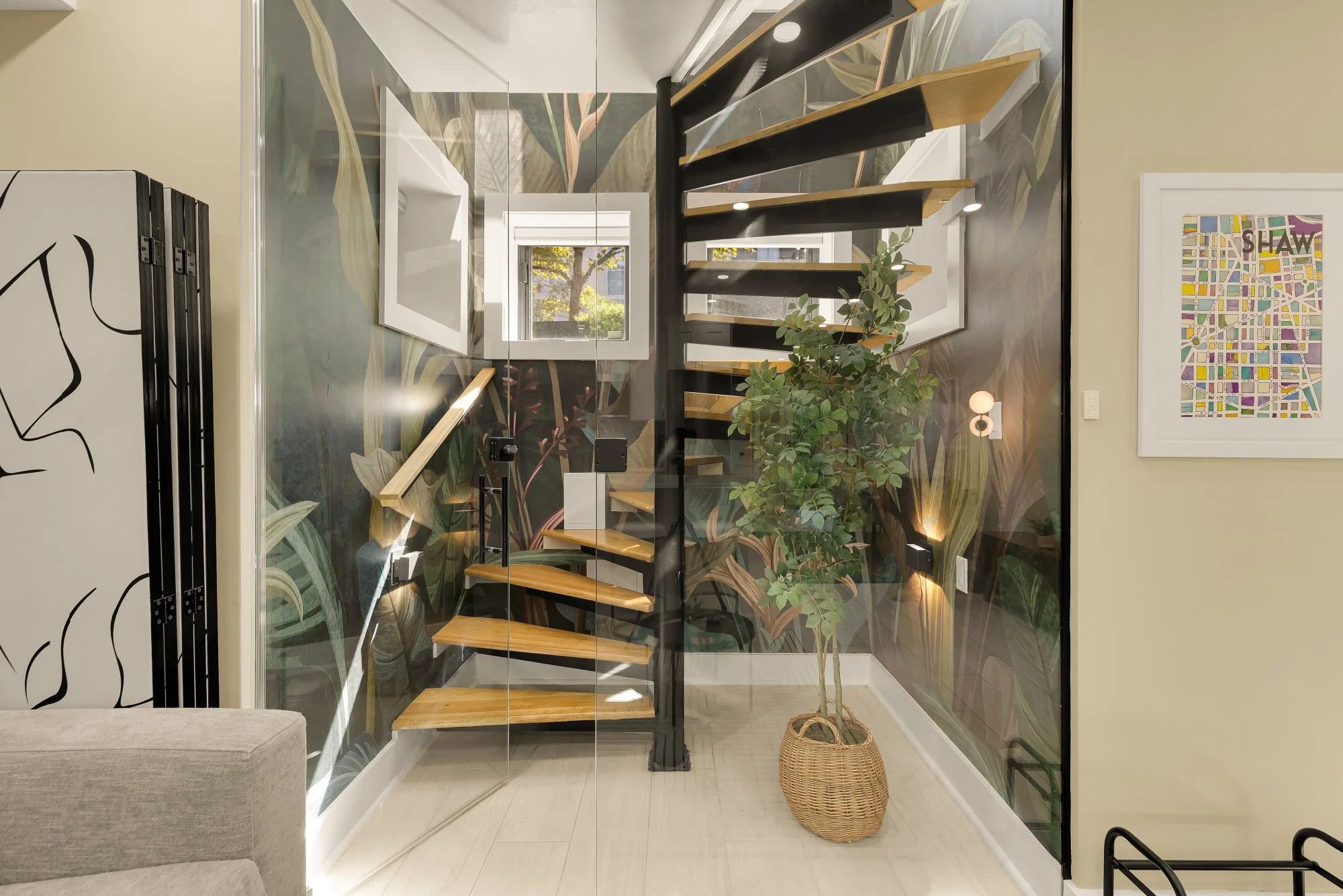 Modern staircase with wooden steps and black metal frame, surrounded by a glass partition with botanical wallpaper, a leafy plant in a woven basket, and artwork on the wall.