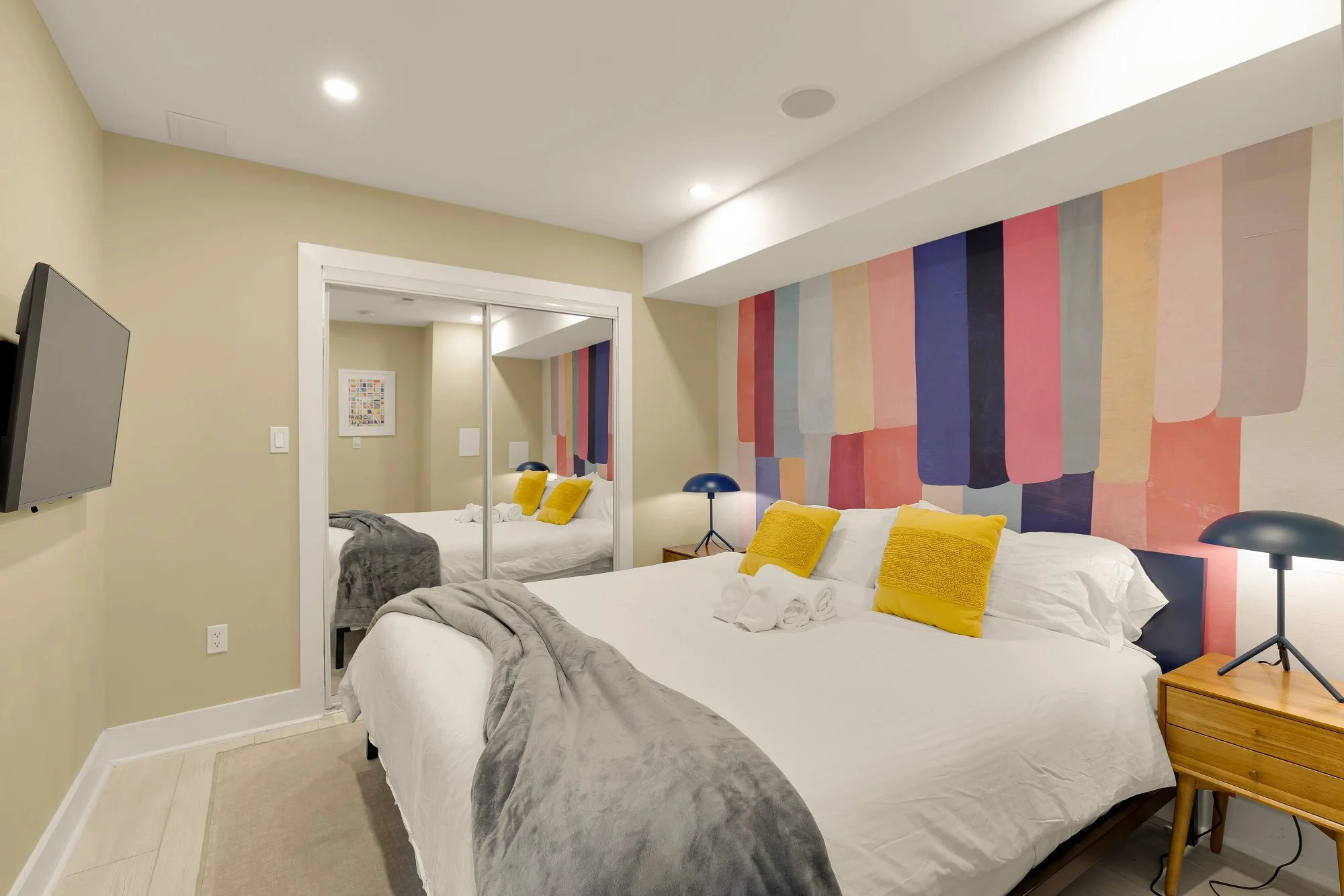 A modern bedroom with two beds, a colorful abstract wall mural, yellow pillows, gray blankets, a wooden nightstand, and a mirror reflecting the room with another bed and artwork on the wall.