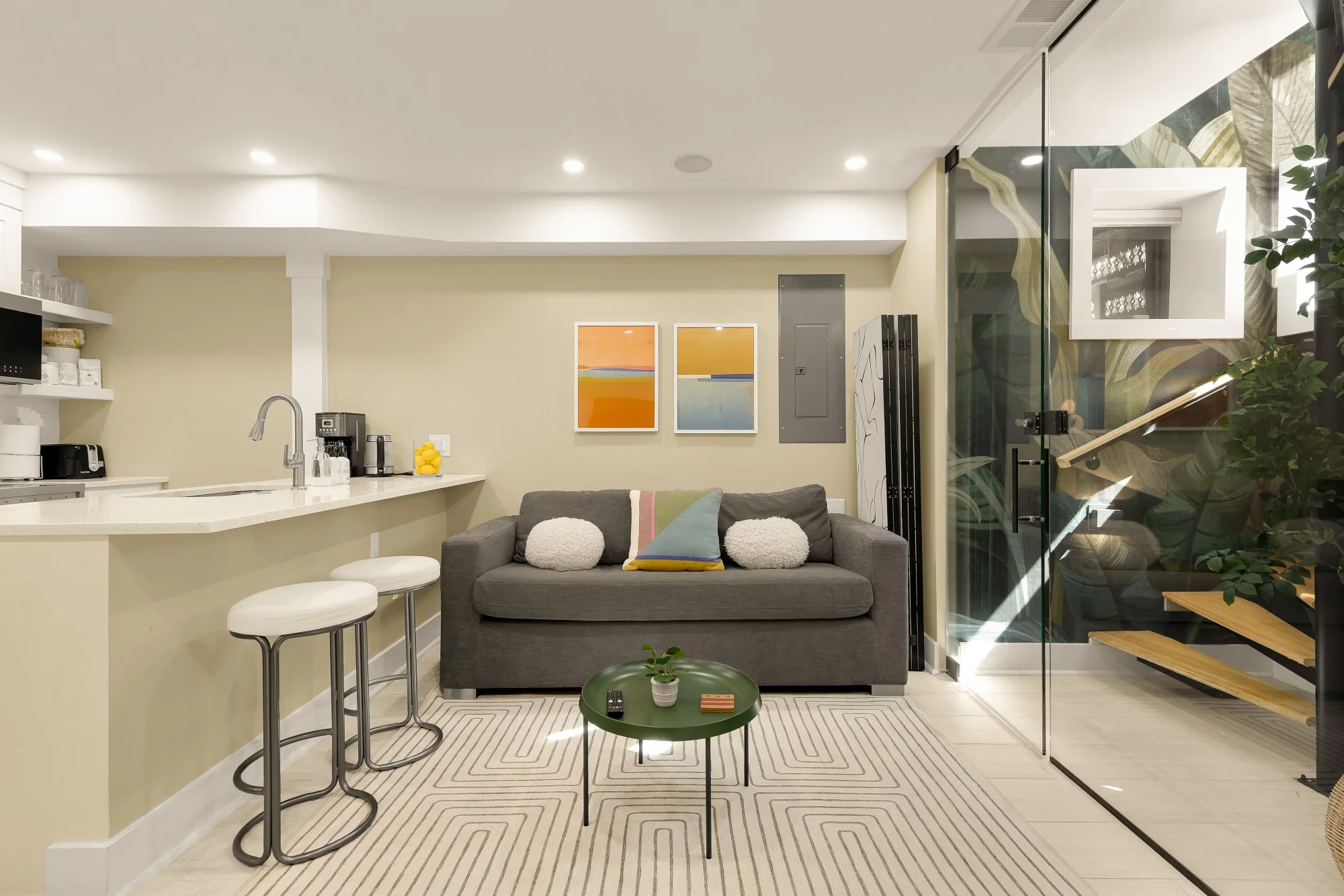 A modern open-concept living space with a kitchen and sitting area. The kitchen has a white countertop with stools, a coffee machine, and a banana fruit holder. The sitting area has a gray sofa with colorful pillows, a small green round table, and ar