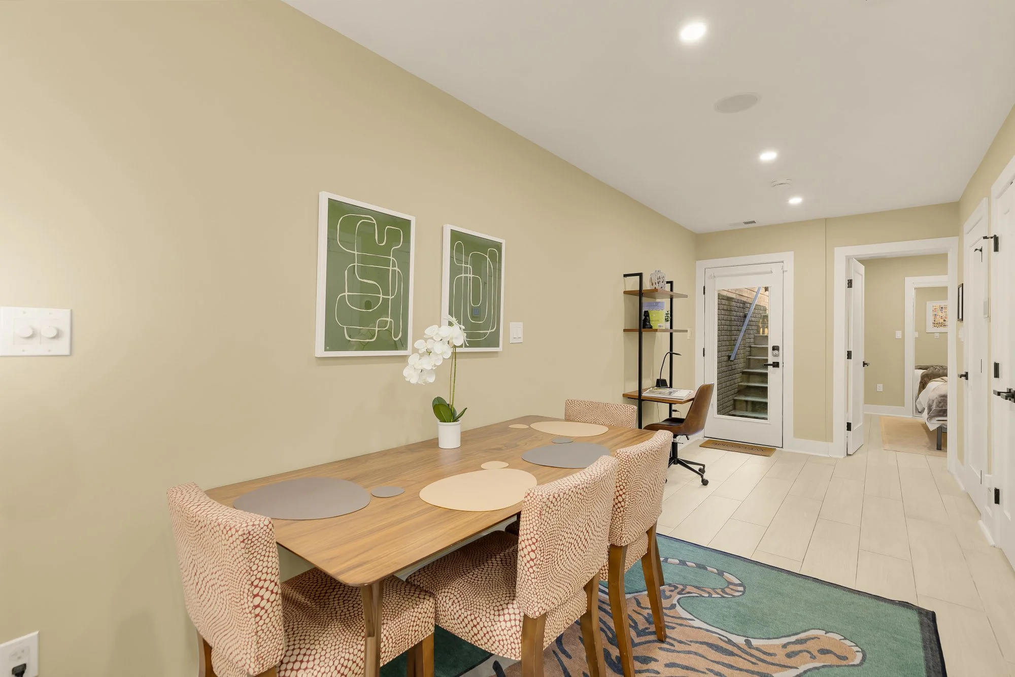 A dining area with a wooden table surrounded by four red patterned chairs, a white orchid in a pot, and wall art with abstract green designs on a light beige wall. Beyond the dining area is a small workspace with a wooden desk, a black office chair, 