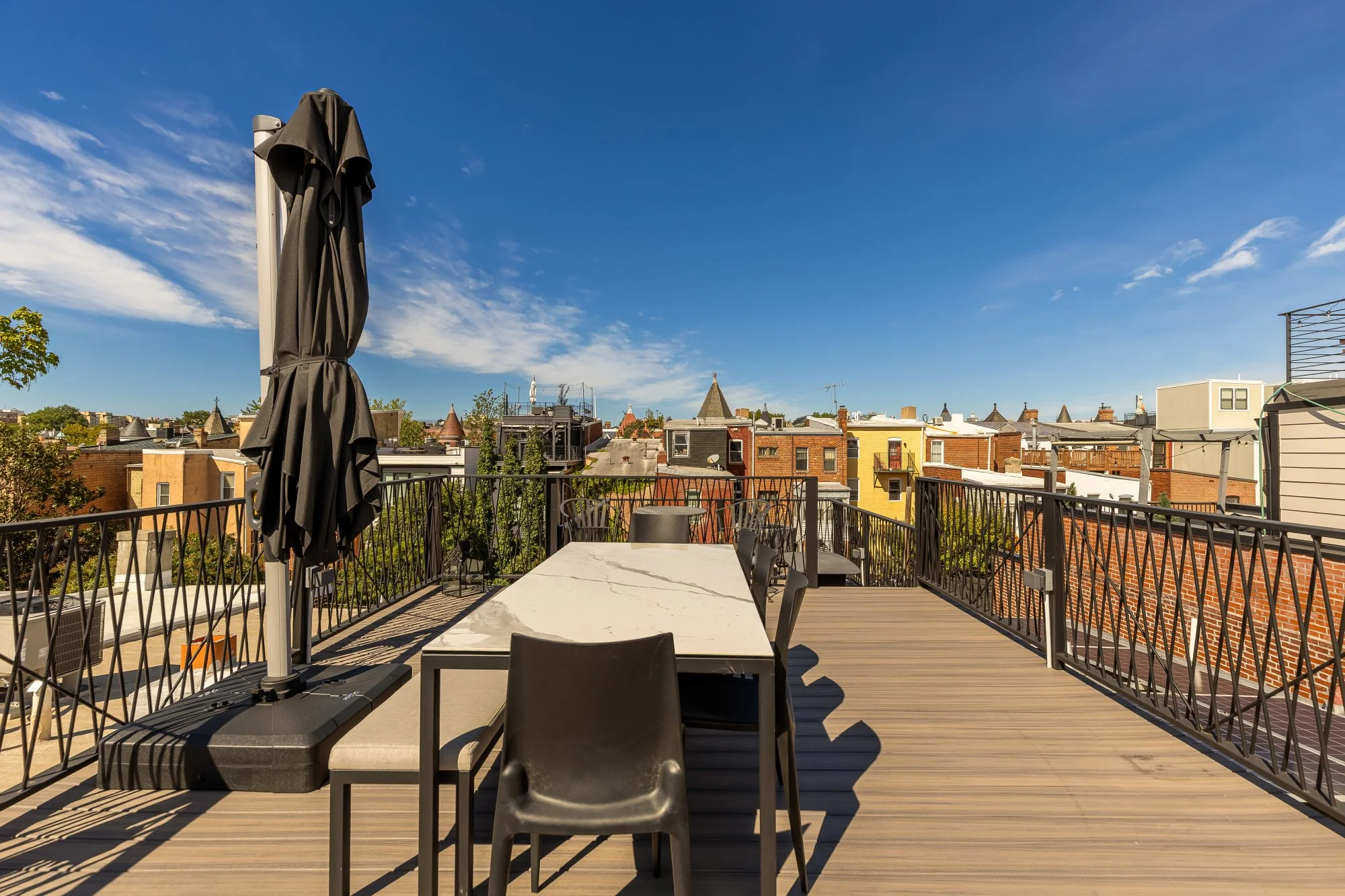A rooftop balcony with a large rectangular table, black chairs, a folded black patio umbrella, and city buildings in the background under a bright blue sky with some clouds.