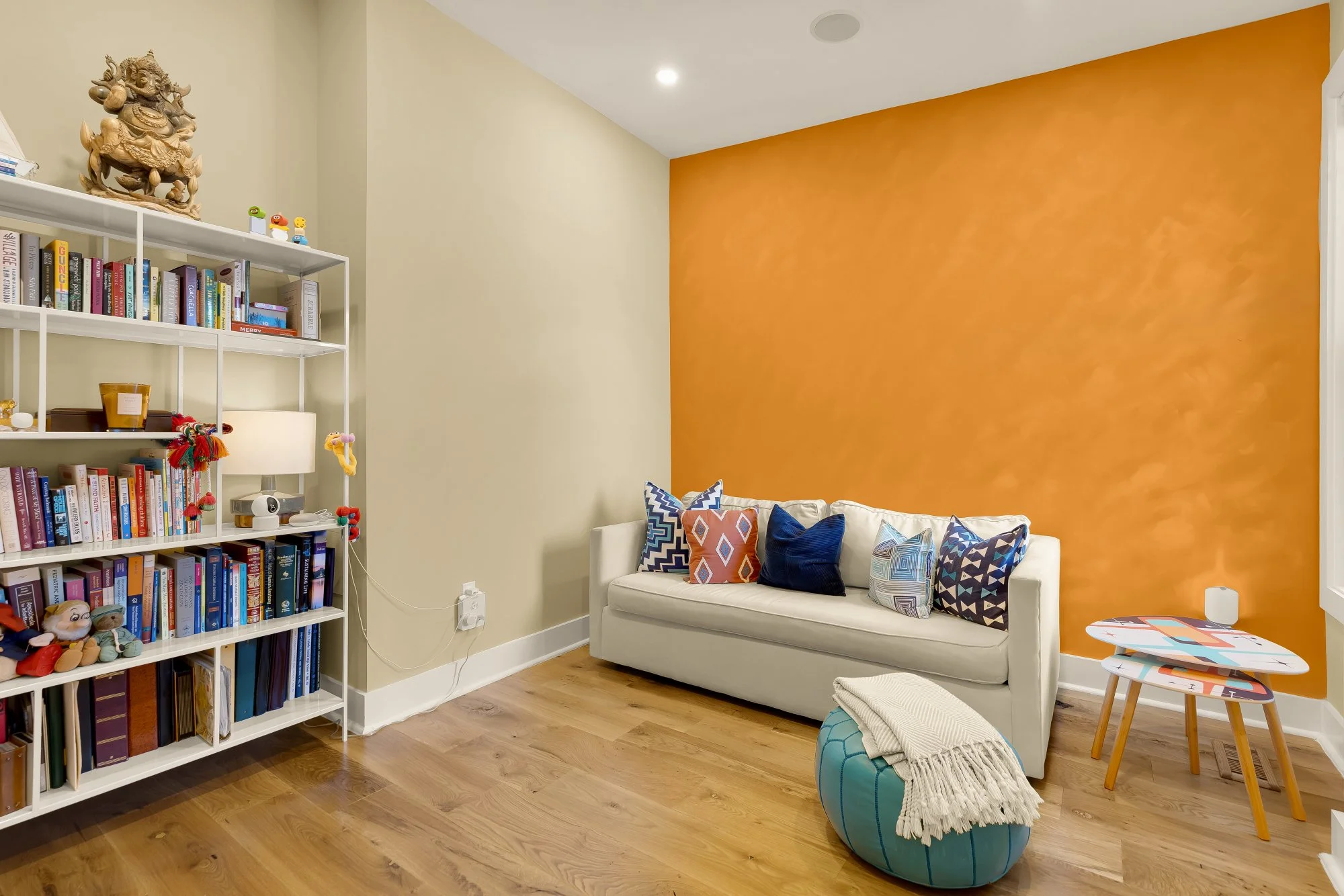 A cozy living room corner with a white sofa adorned with colorful patterned pillows, a large orange accent wall, a white bookshelf filled with books and decorations, a small round table with children's play items, and a beanbag ottoman with a throw b