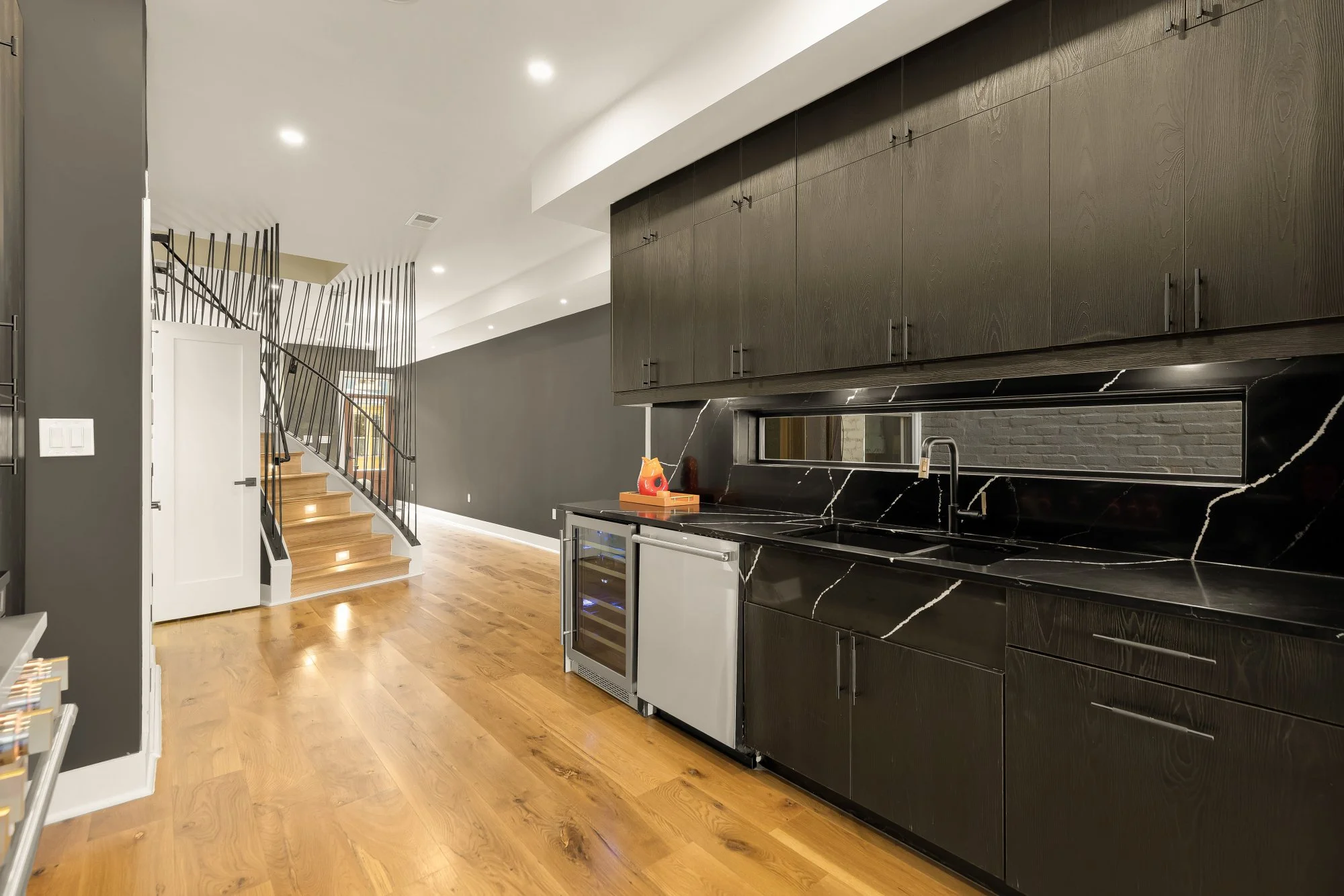 Modern kitchen with black marble countertops, dark wooden cabinets, stainless steel appliances, and hardwood flooring. An open staircase with black metal railing is visible in the background.
