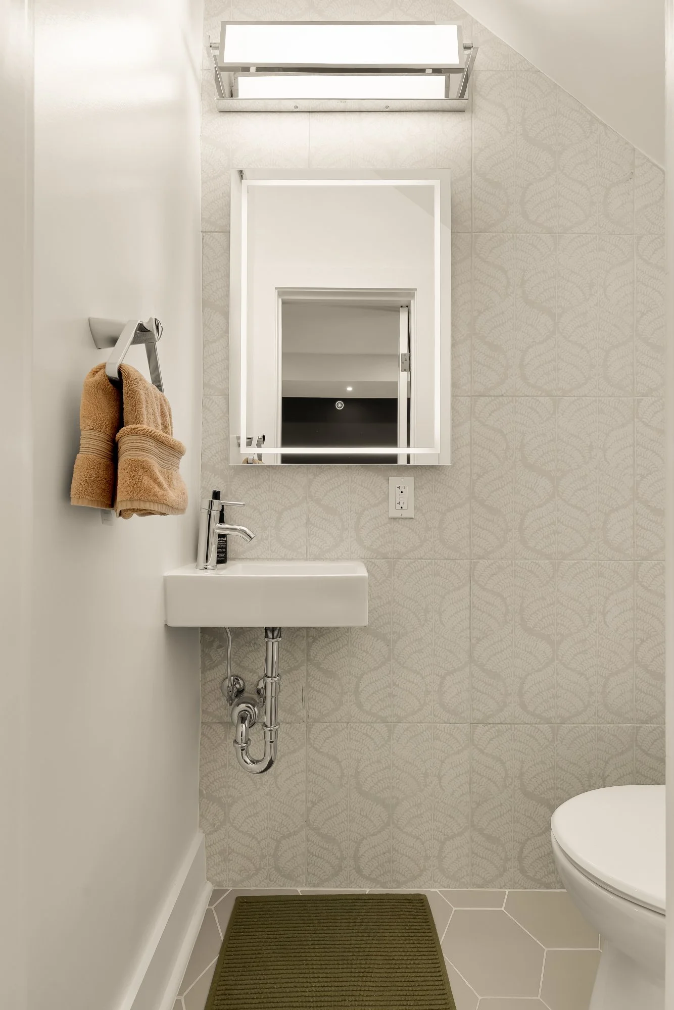 A modern bathroom with a small sink, a mirror, a towel rack with two towels, an electrical outlet, and part of a toilet. The walls have a light pattern, and a brown striped rug is on the floor.