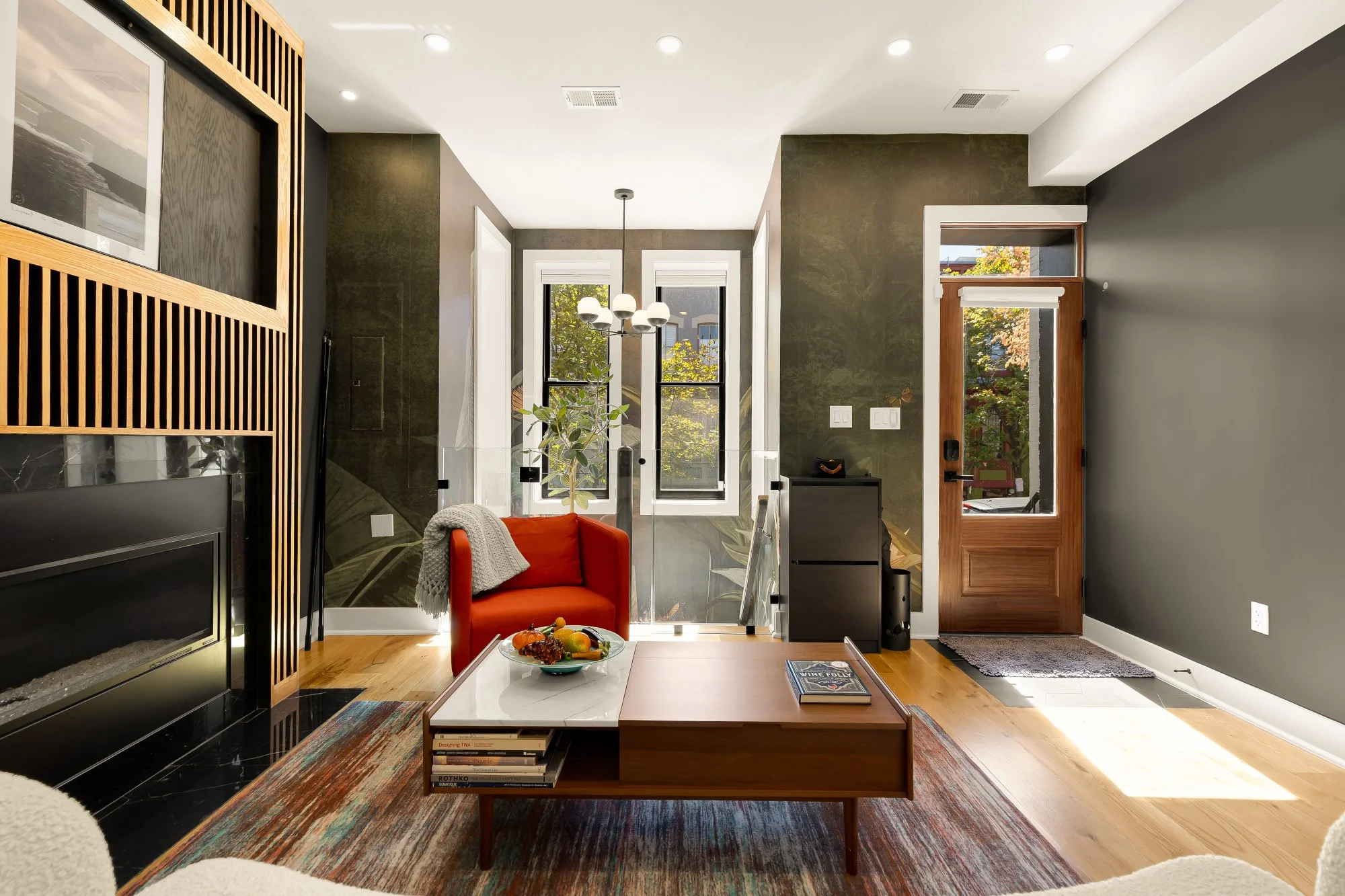 Modern living room with a white ceiling, dark walls, and large windows. A hanging light fixture with multiple bulbs is centered in the room. There is a red armchair with a throw blanket, a dog gate, a black side table, and a wooden door with glass. A