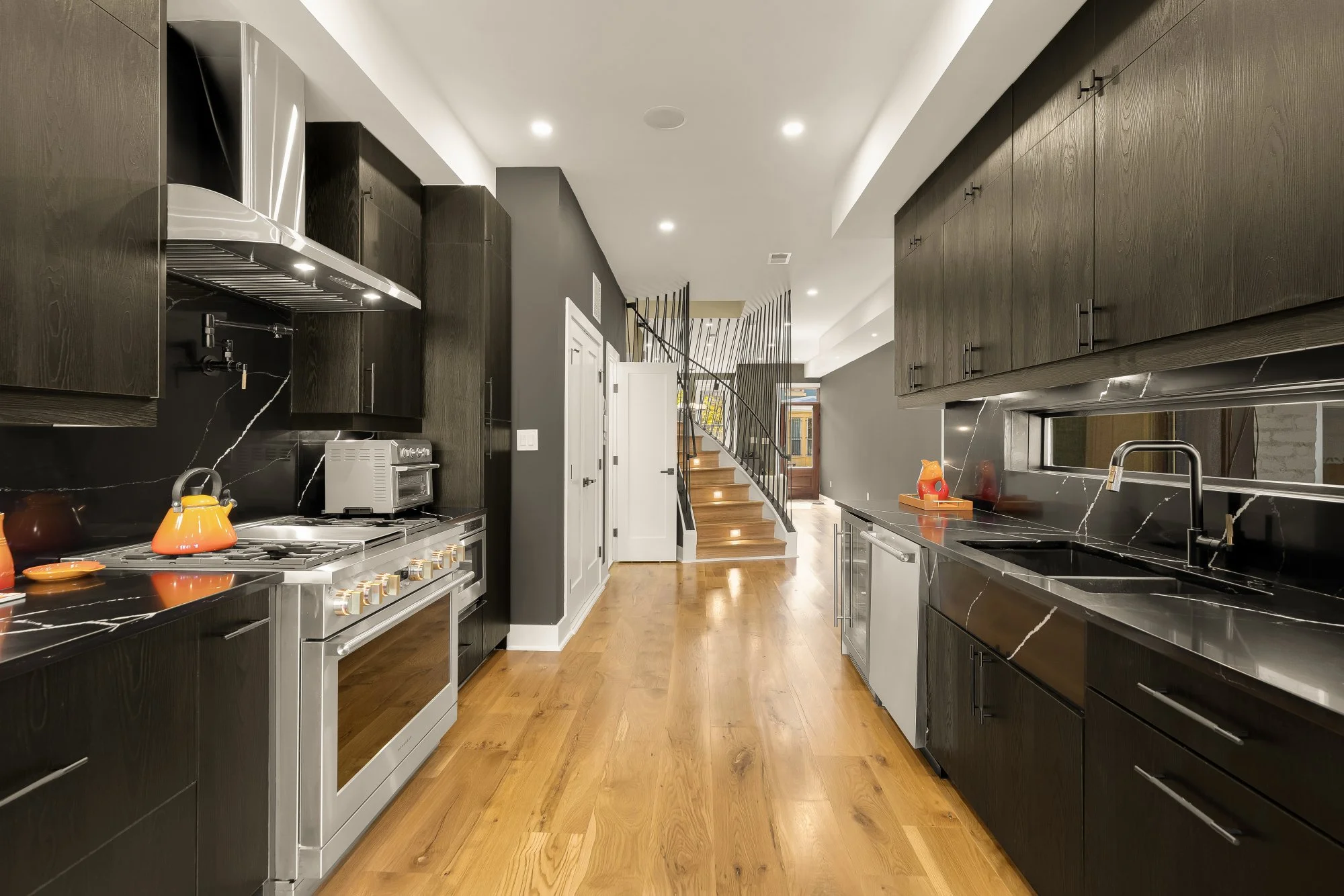 Modern kitchen with black cabinets, marble countertops, stainless steel appliances, wooden floors, and a stairway leading to an upper level.