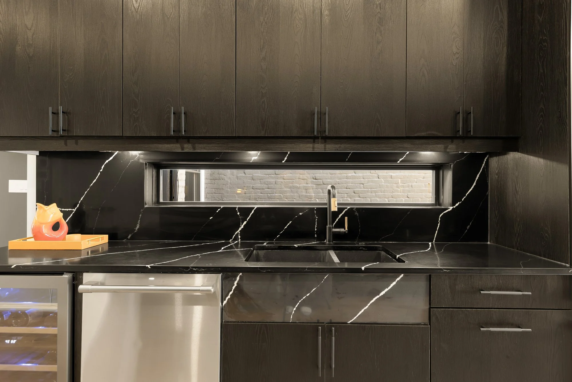 Modern kitchen with black marble countertop and backsplash featuring white veining, dark wooden upper cabinets with silver handles, a small window above the sink, a yellow tray with a decorative orange and yellow ceramic jug on the left, and a stainl